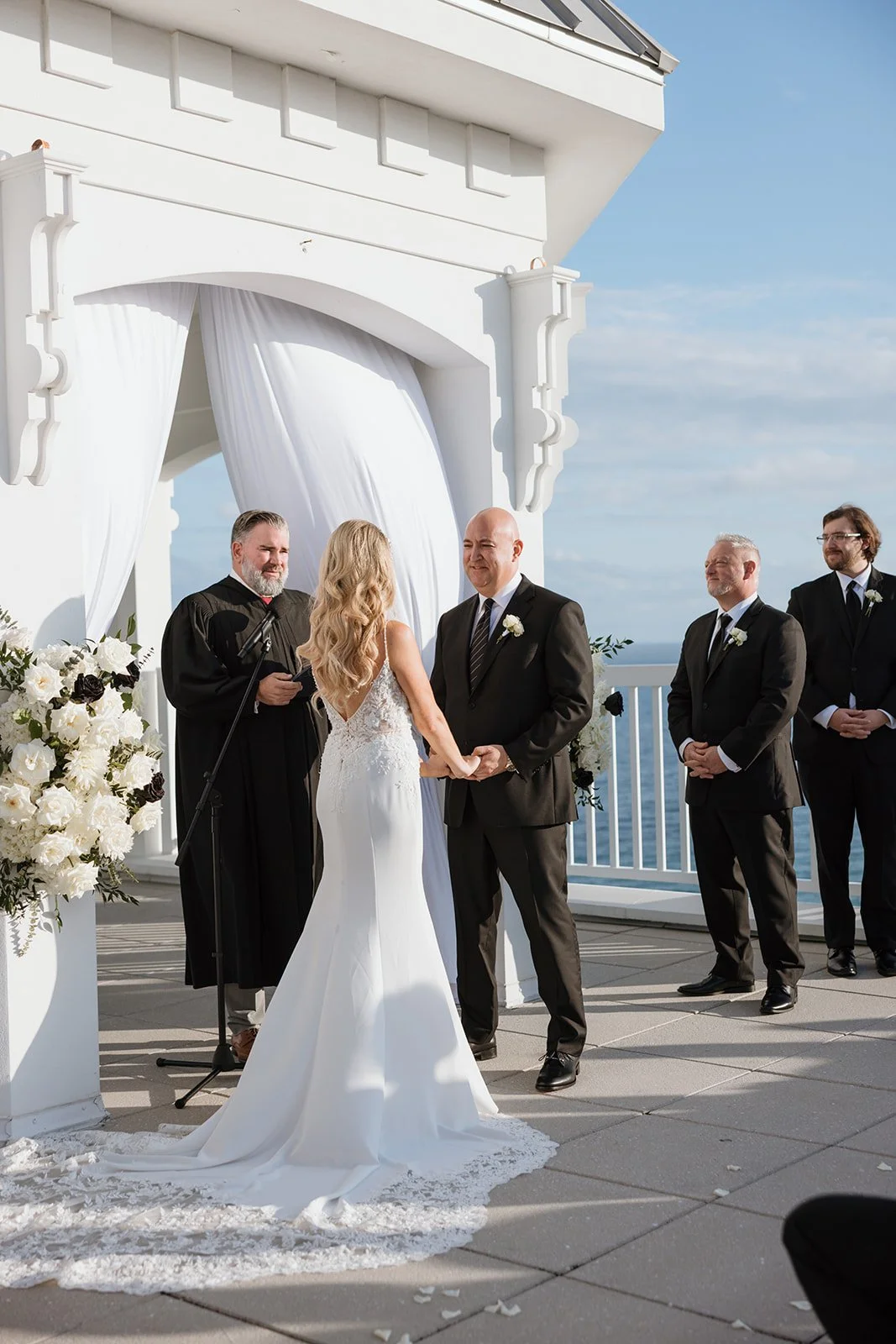 A couple getting married at an outdoor ceremony overlooking the ocean, with three groomsmen, a woman officiant, and a decorative white archway with flowers.