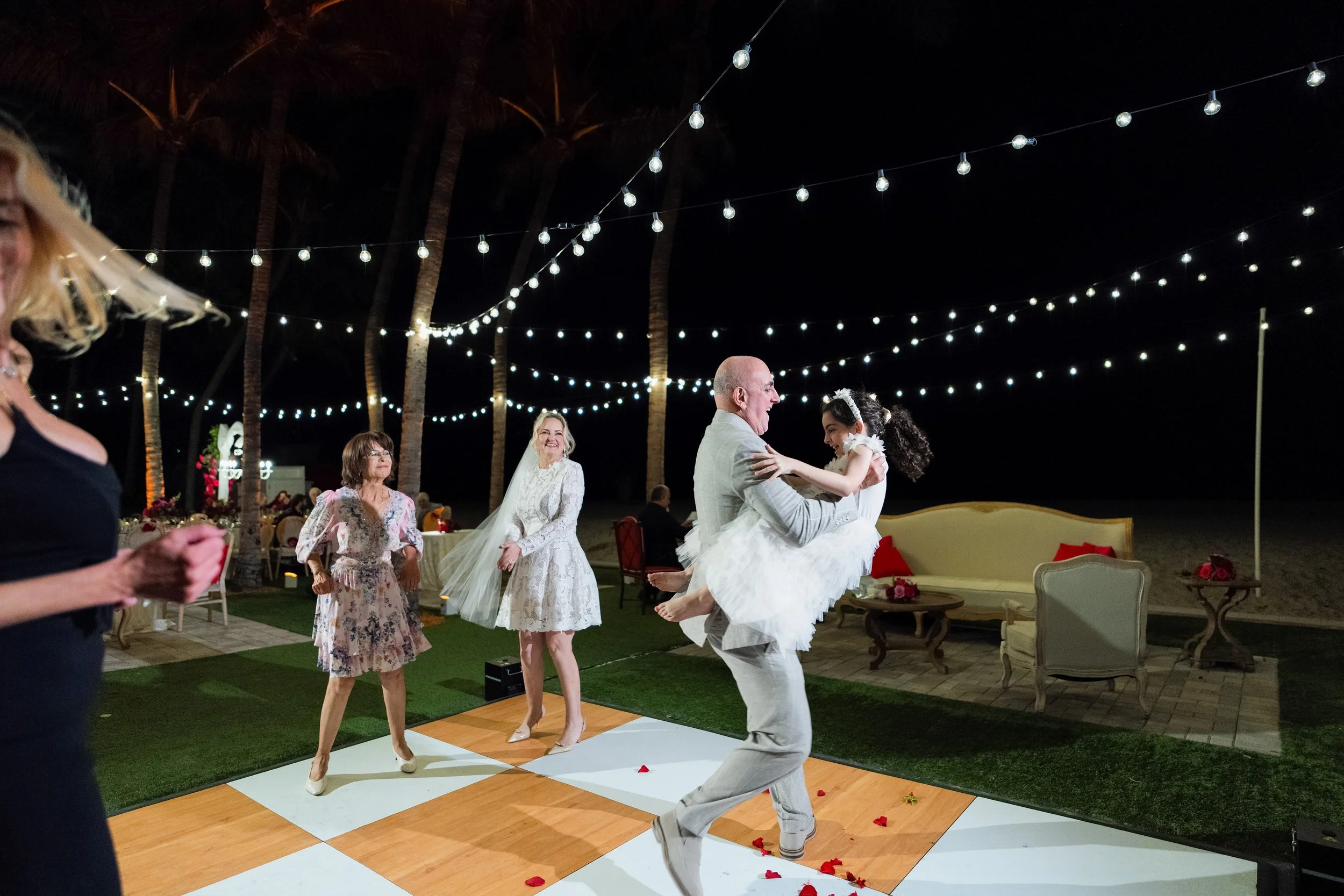 A nighttime outdoor wedding reception scene with string lights, palm trees, and a dance floor, where a man is lifting a young girl in a white dress and a girl with a veil, all smiling.