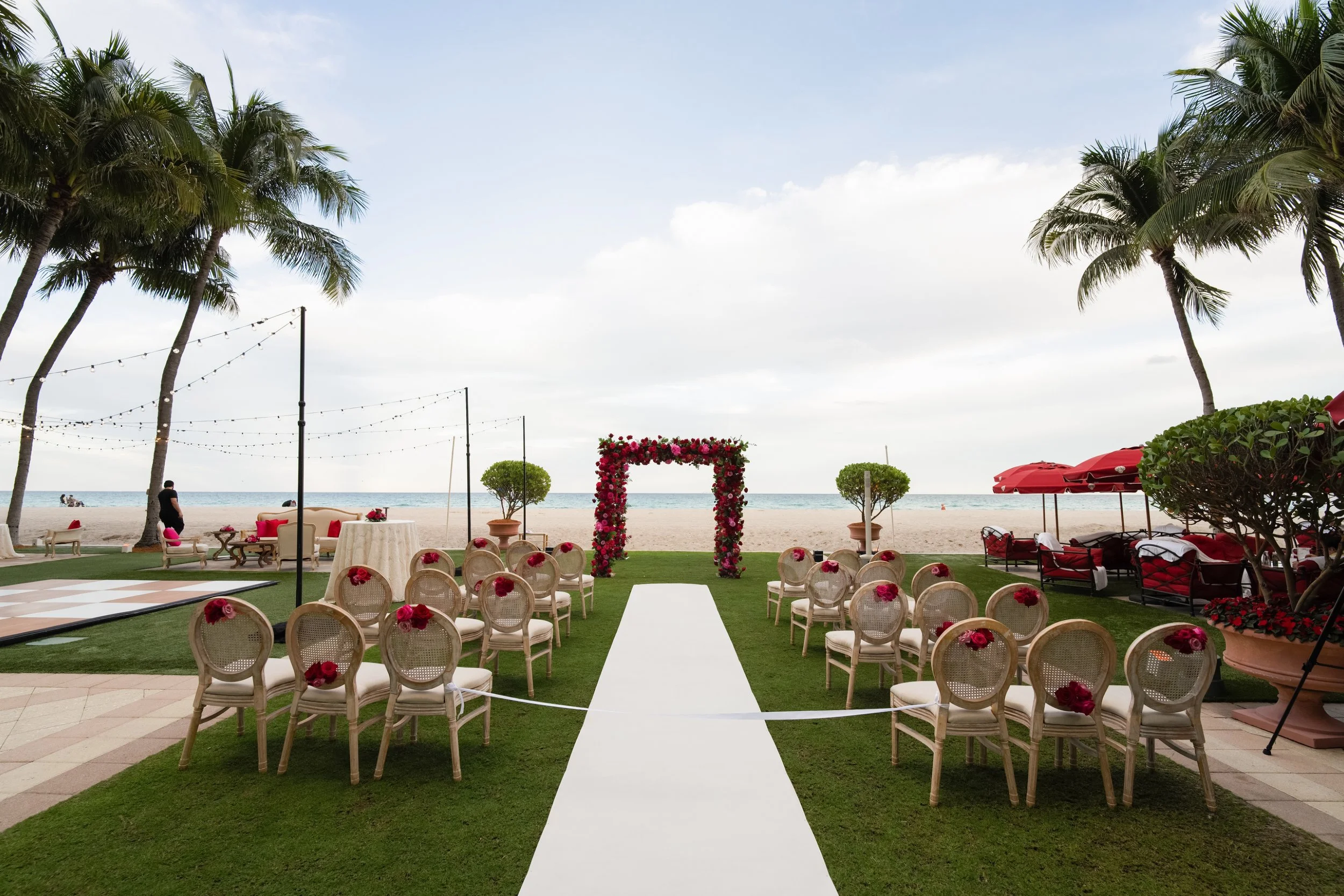 Beachside wedding ceremony setup with white chairs decorated with red flowers, a floral arch, and a view of the ocean with palm trees and seating areas under red umbrellas.