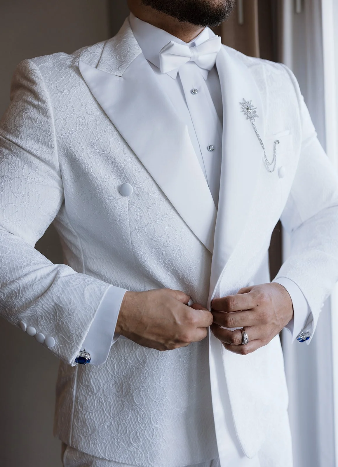 Man in a white tuxedo with a textured pattern, adjusting the jacket. He is wearing a bow tie, a brooch pin, and a wedding ring.