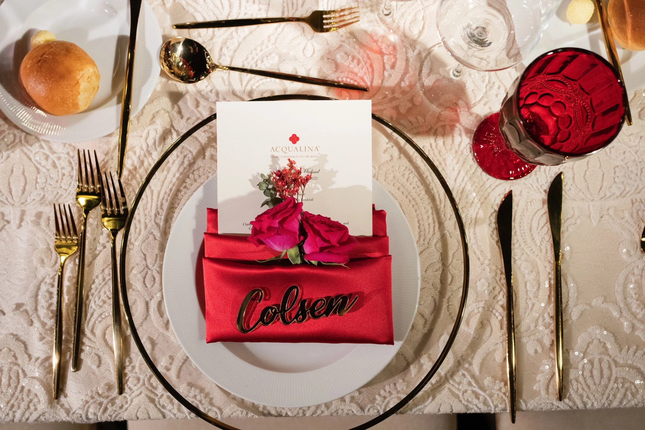 Elegant table setting with a white plate, gold utensils, a red napkin with the name 'Colsen,' pink flower, invitation card, wine glass, and red goblet on a lace tablecloth.