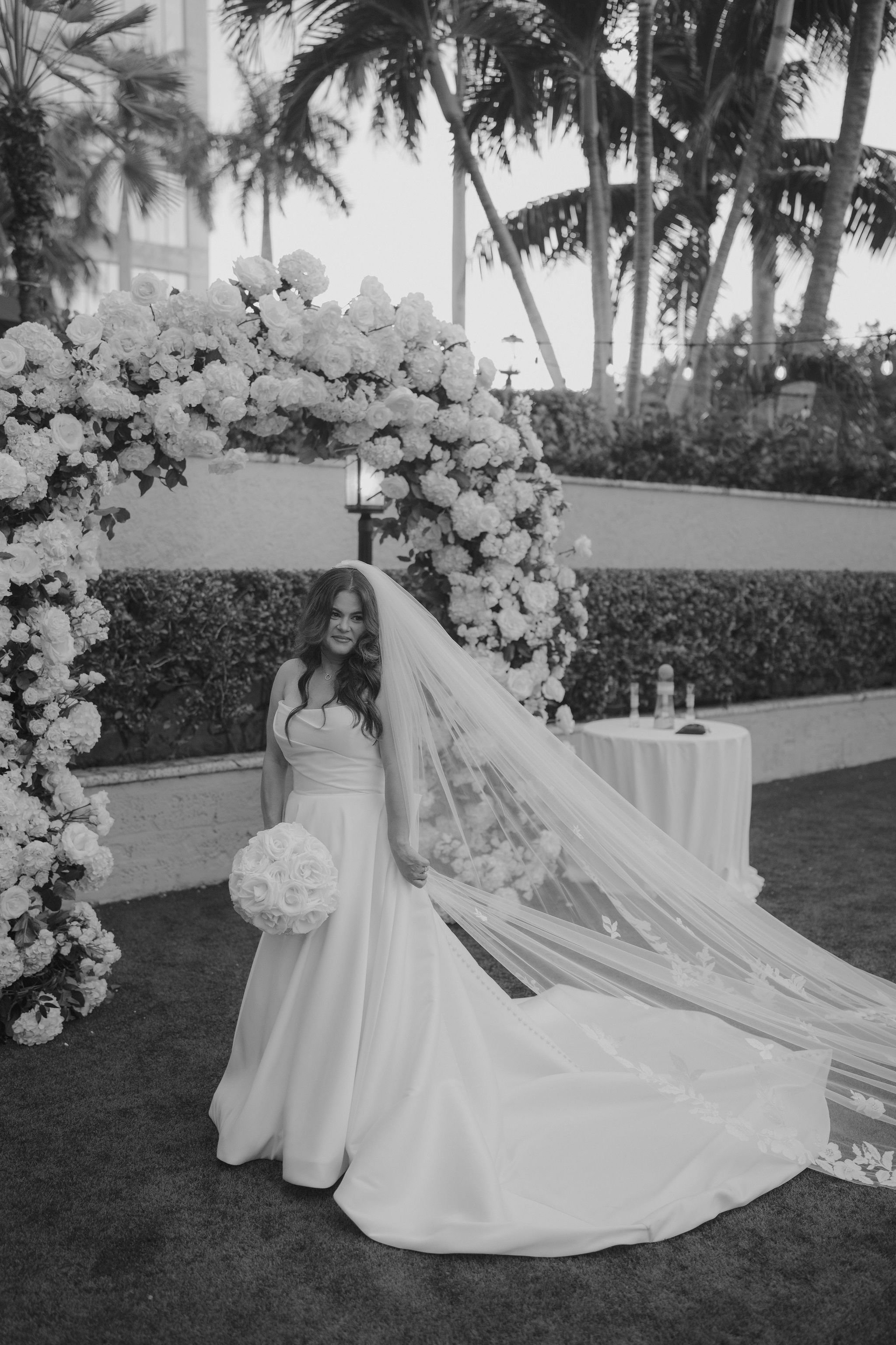 A bride in a wedding dress holding a bouquet of roses, standing in front of a floral arch outdoors.