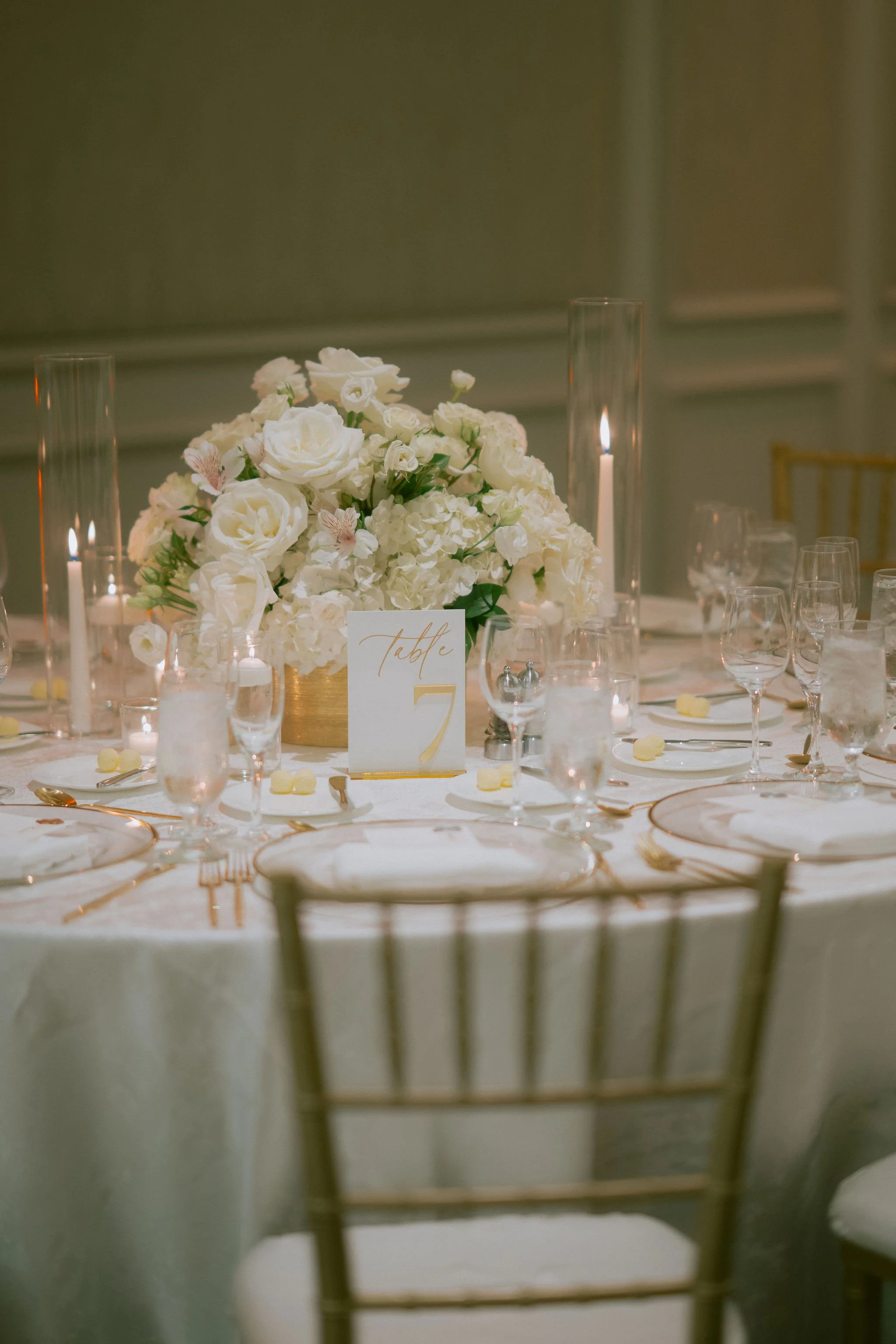 Elegant wedding reception table decorated with a large floral centerpiece featuring white roses and hydrangeas, surrounded by glassware, gold cutlery, and a table number card indicating 'Table 7'. Candles in tall glass holders add to the sophisticate