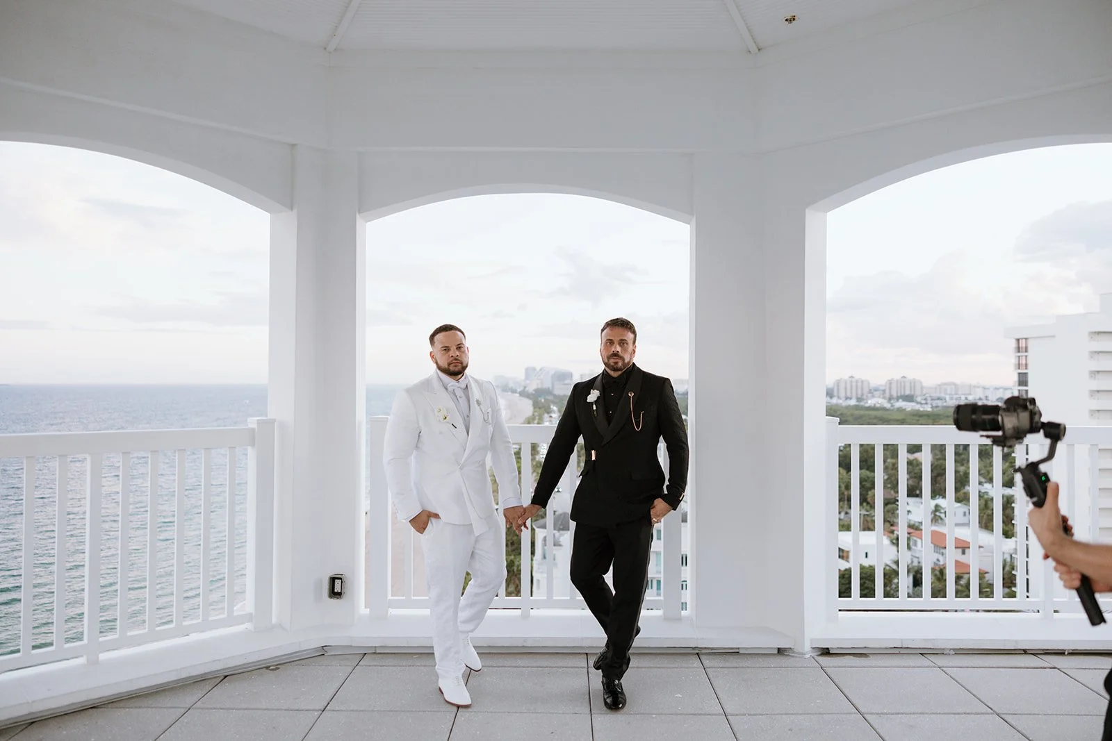 Two men dressed in formal suits holding hands on a balcony with a city skyline and water in the background, while a person takes a photo with a camera.