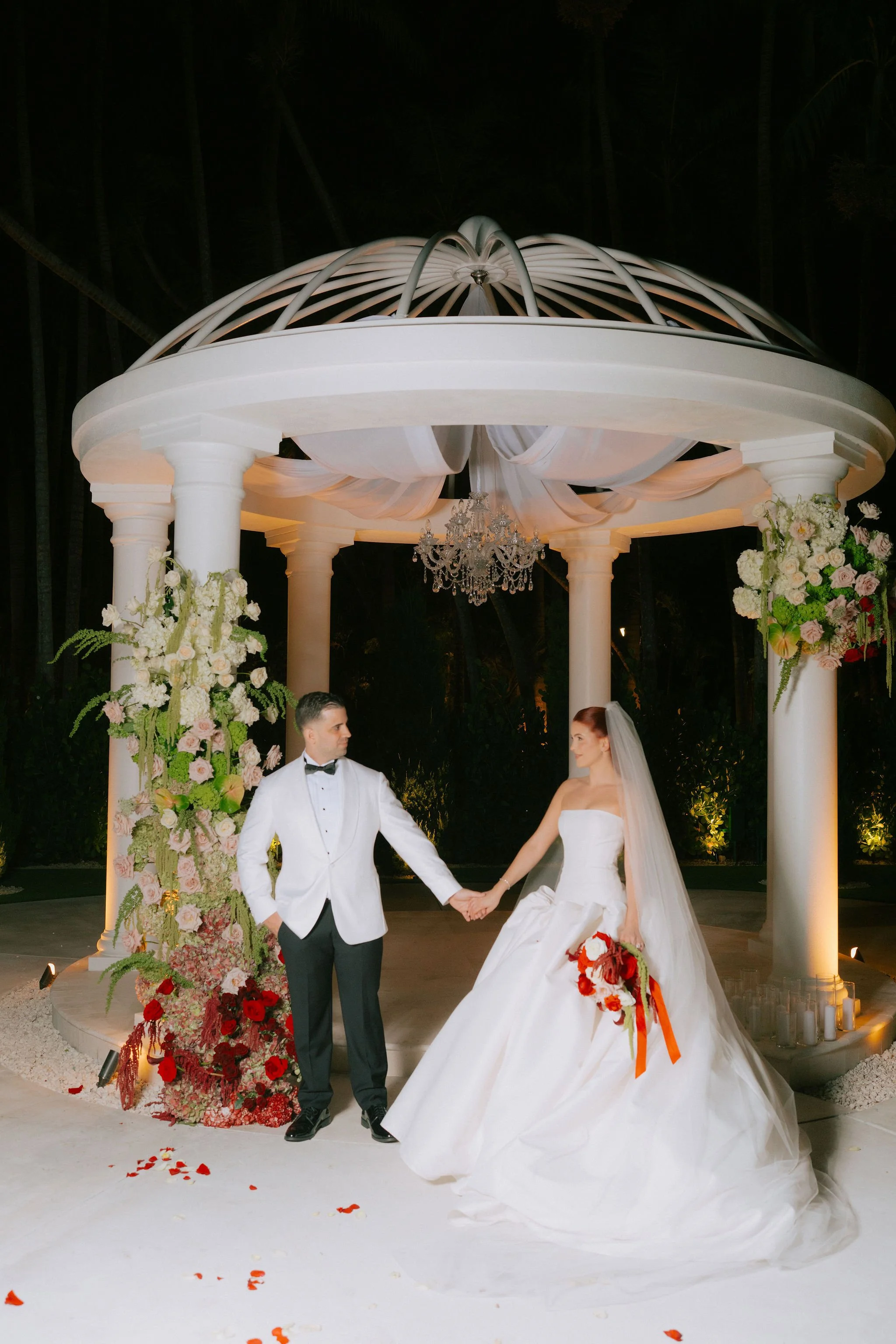 A bride and groom holding hands under a decorative gazebo at their wedding ceremony. The bride is wearing a white gown with a veil, and the groom is dressed in a white tuxedo jacket and black pants. There are floral arrangements on the gazebo columns