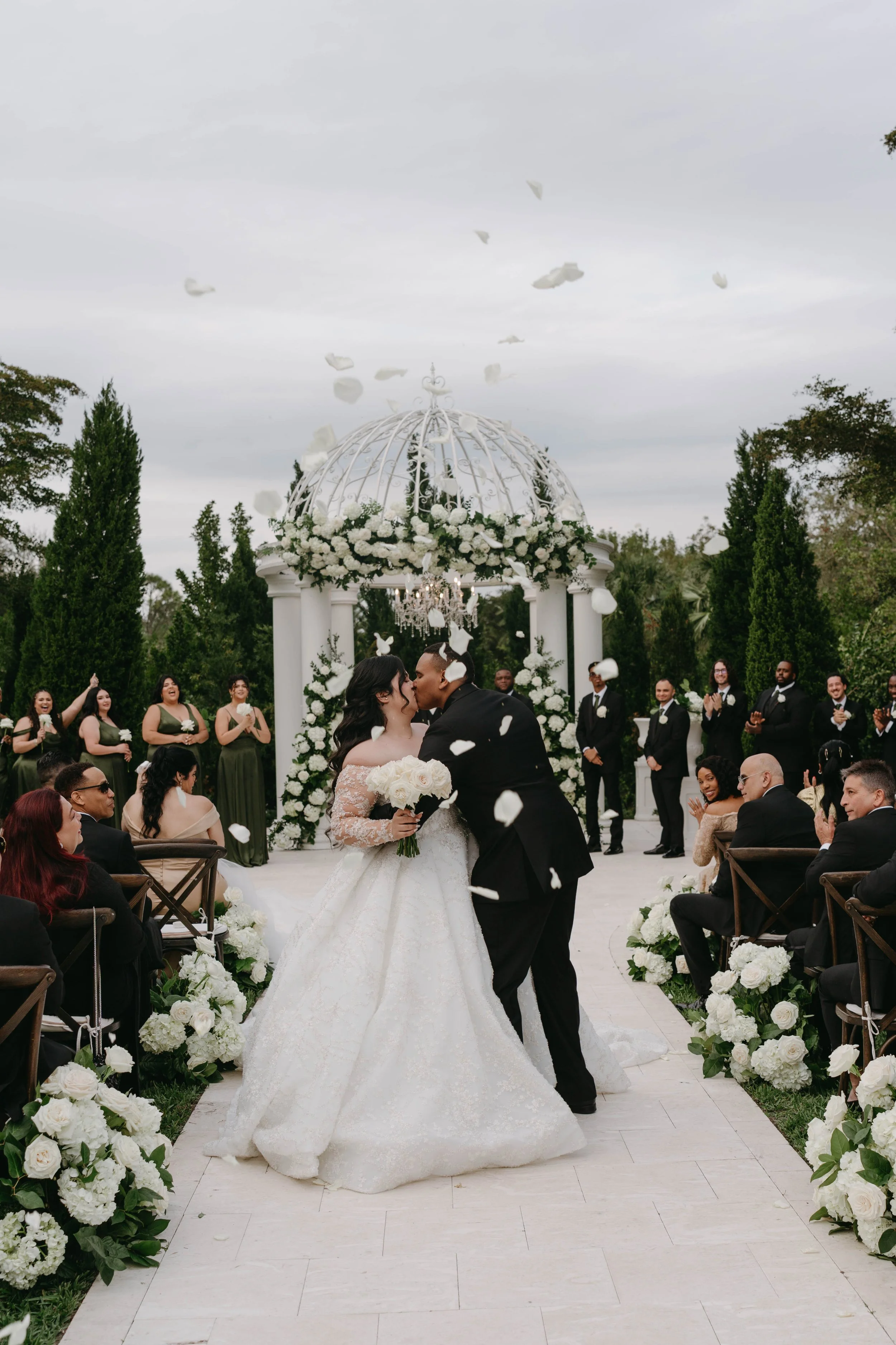 A wedding ceremony outdoors with a bride and groom kissing, surrounded by guests and floral decorations, with white flower petals being tossed in the air.