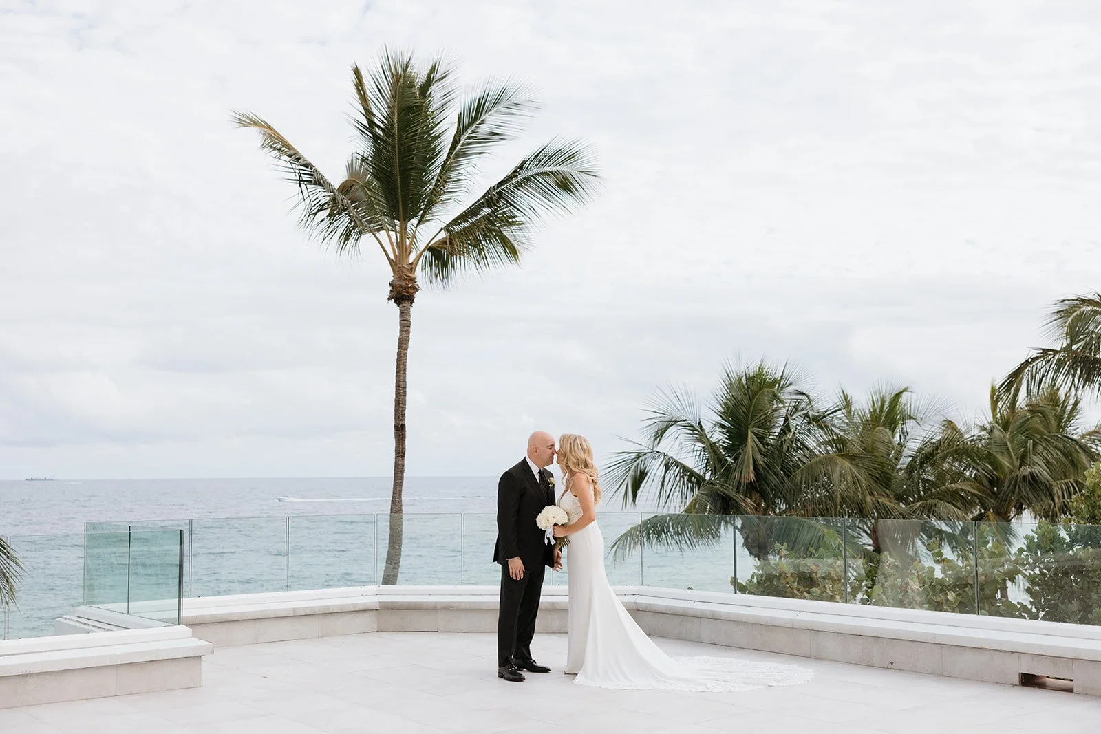 Bride and groom kissing in elegant interior with staircase and grand piano, wedding attire, floral bouquet, ceiling chandelier.