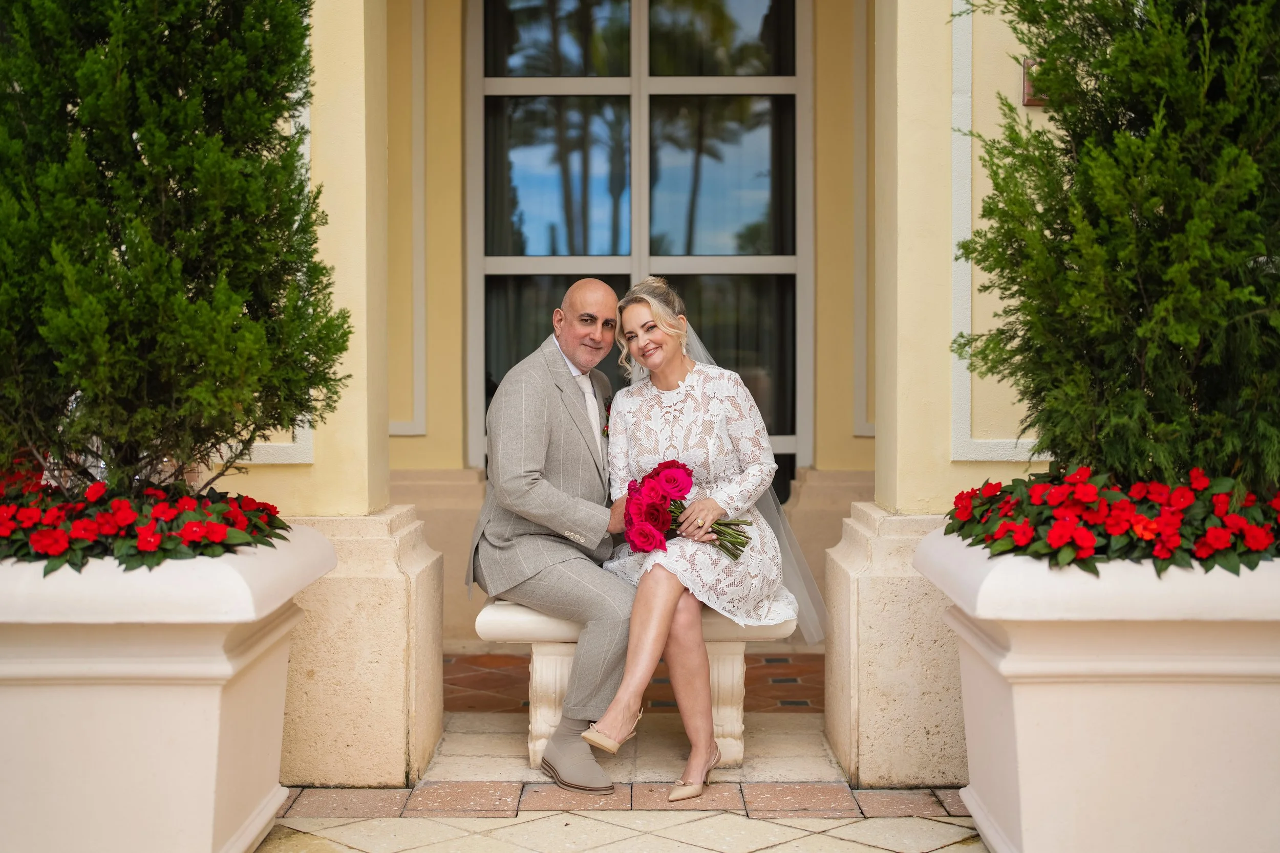A bride and groom sitting on a white bench outside a building, smiling and holding red roses. There are two large planters with red flowers and green bushes on either side of them.