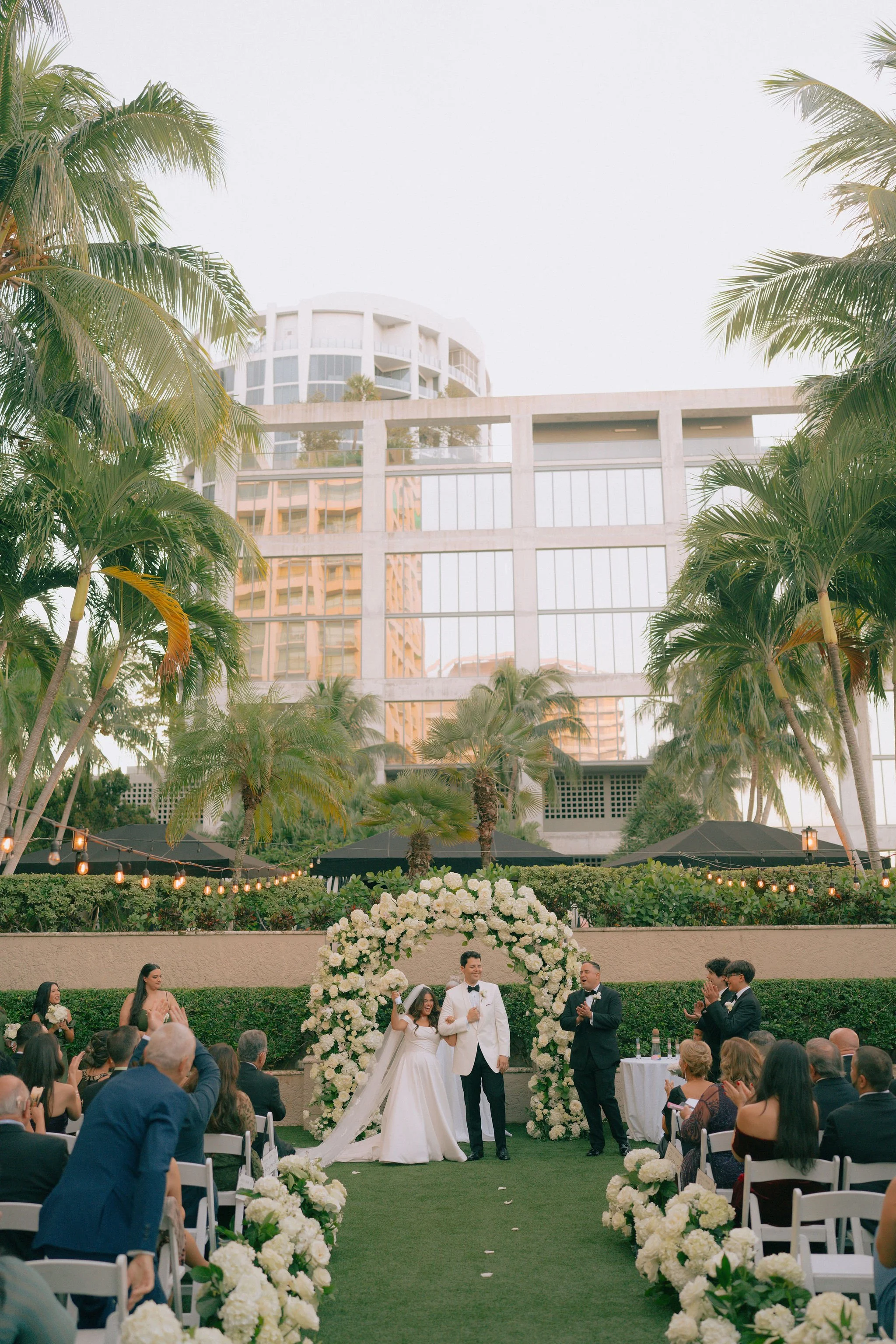Outdoor wedding ceremony with a bride in a white gown and a groom in a white tuxedo under a floral arch, surrounded by guests and lush tropical trees, with tall modern buildings in the background.