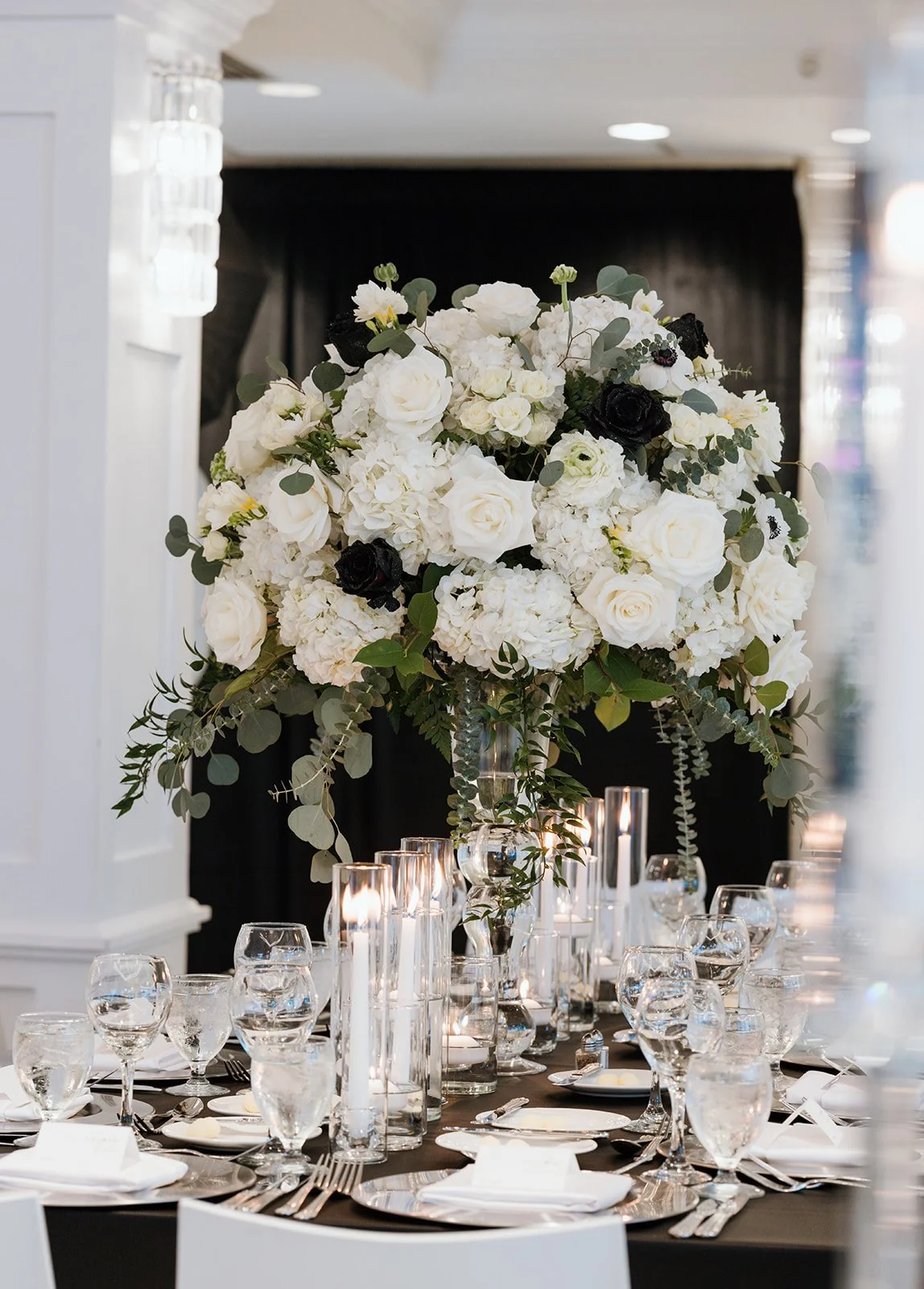 Elegant banquet table with a large floral centerpiece of white roses, hydrangeas, and greenery, surrounded by glasses, plates, and silverware, on a black tablecloth.