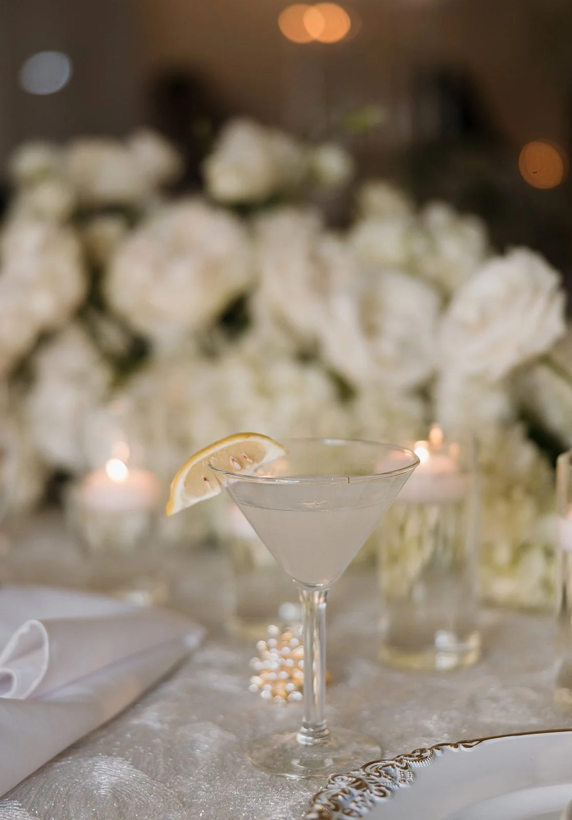 A clear cocktail glass with a lemon wedge on the rim, set on a decorated table with white flowers and candles in the background.