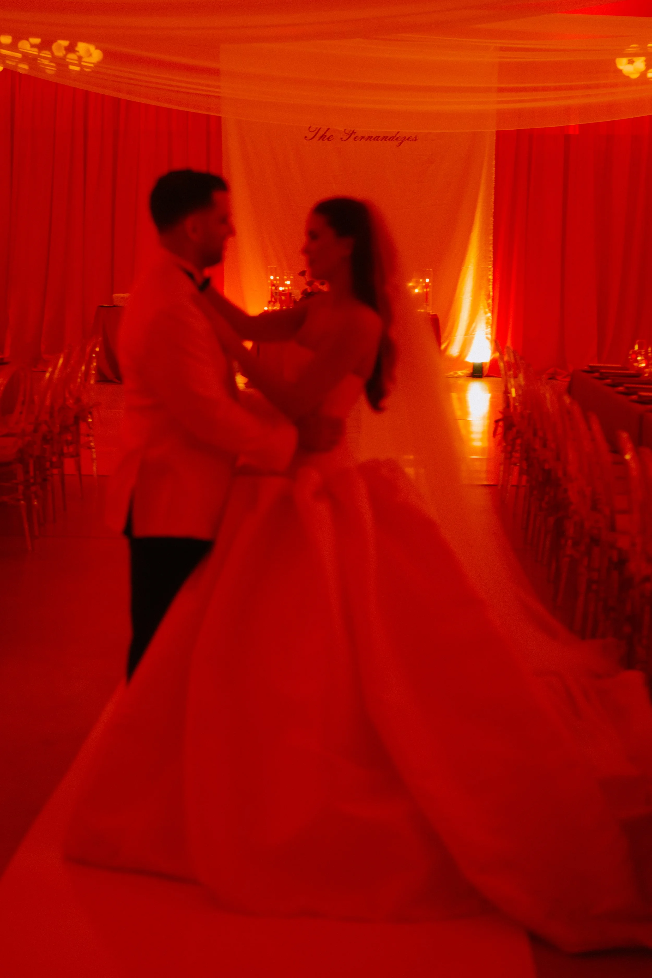 A bride and groom dancing in a warmly lit reception hall, with a backdrop reading 'The Fernandezes'.