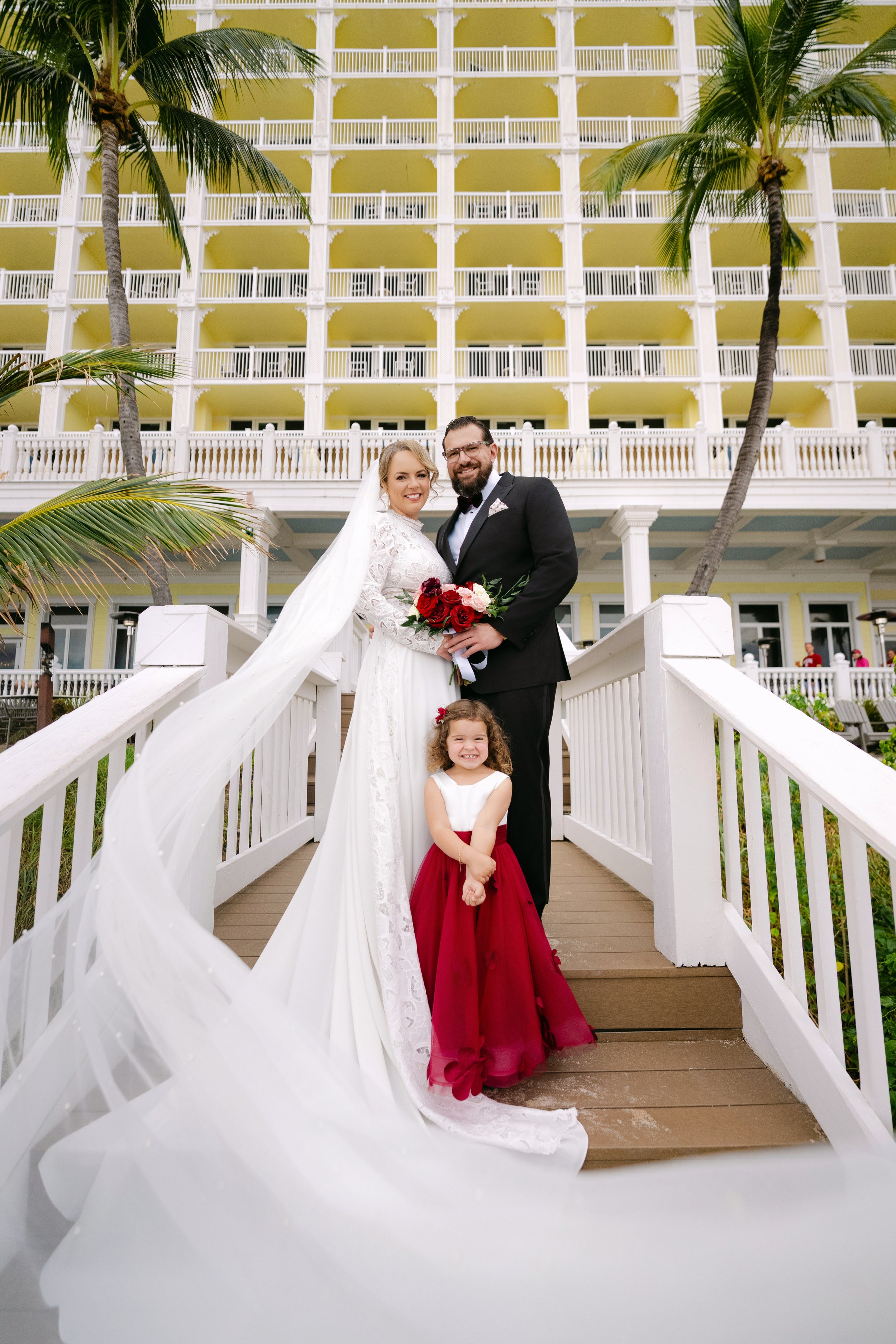 A bride and groom pose on a staircase outside a yellow resort building with white balconies. The bride wears a white wedding dress with a veil and holds a bouquet of red and white flowers. The groom wears a black tuxedo. A young girl in a red dress s