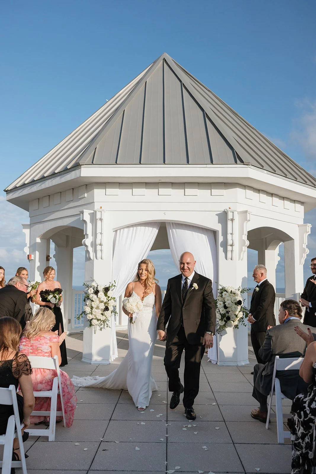 A bride and groom walk down the aisle after their wedding ceremony at an outdoor venue with a white gazebo, surrounded by guests clapping and taking photos.