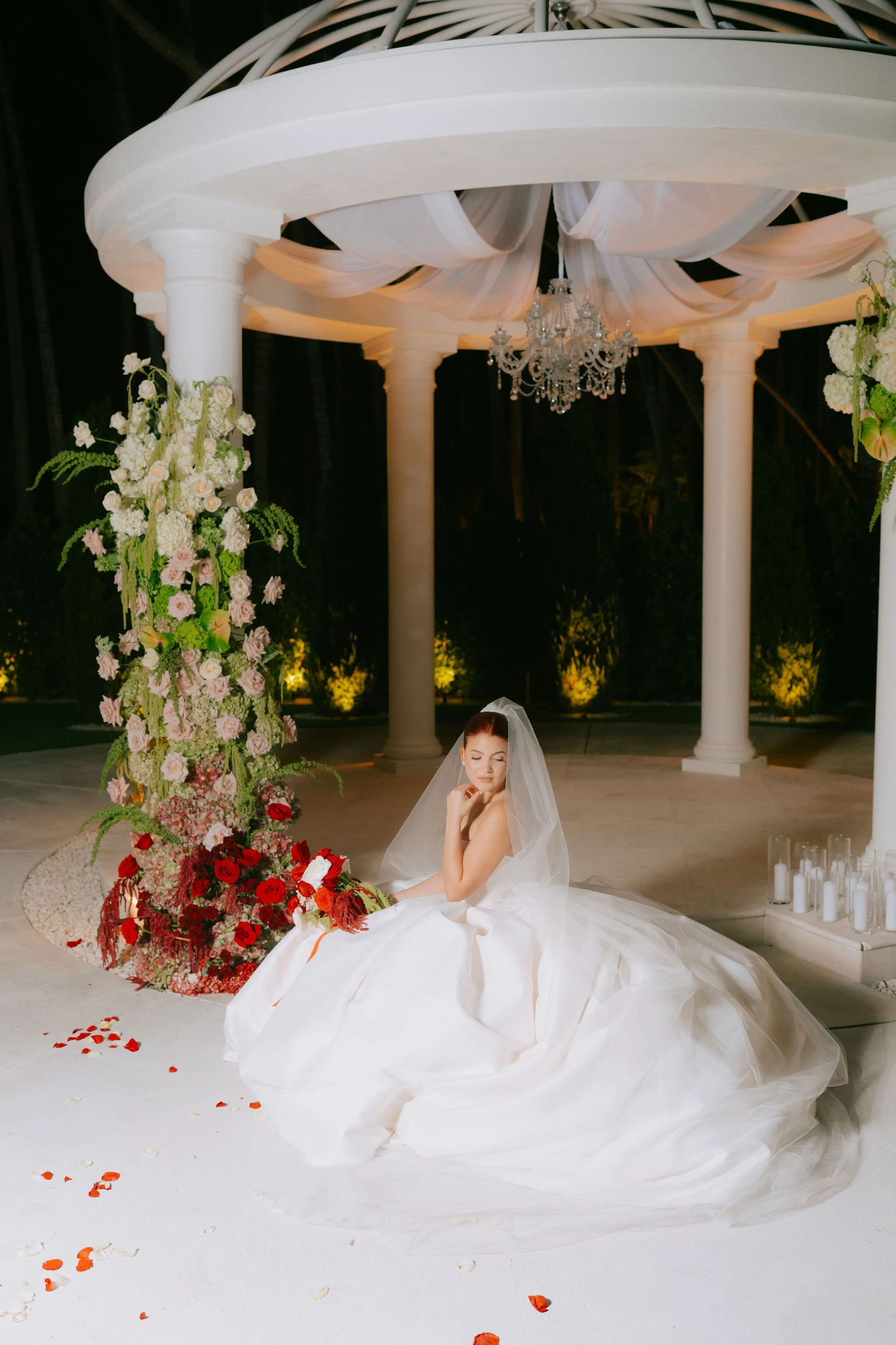 A bride in a white wedding gown and veil sitting on the floor next to a large floral arrangement with pink, red, and white flowers, under a white pavilion with columns and draped fabric, illuminated at night.