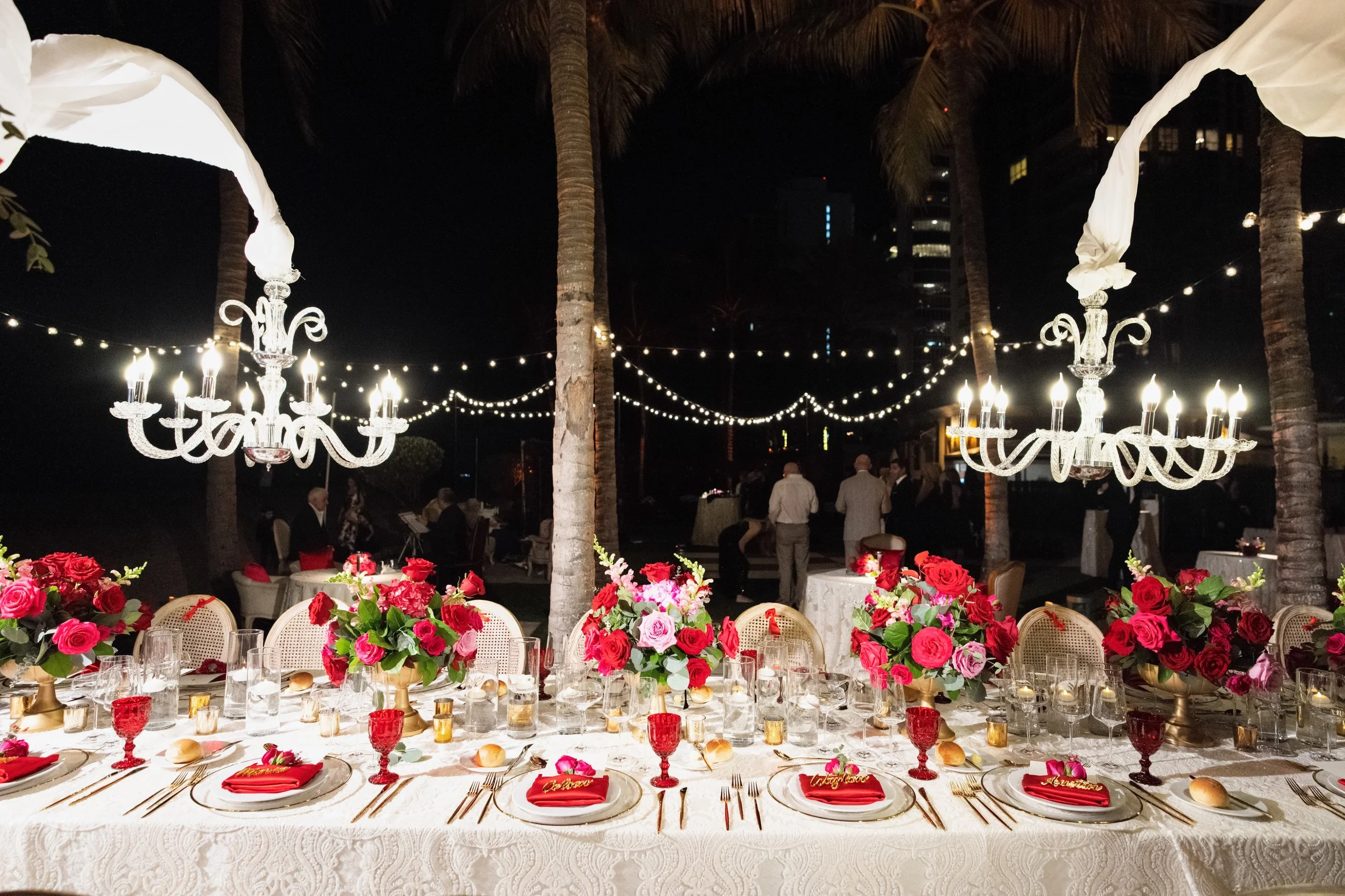 A decorated outdoor dining setup at night with a long table, chandeliers, floral arrangements, and string lights.