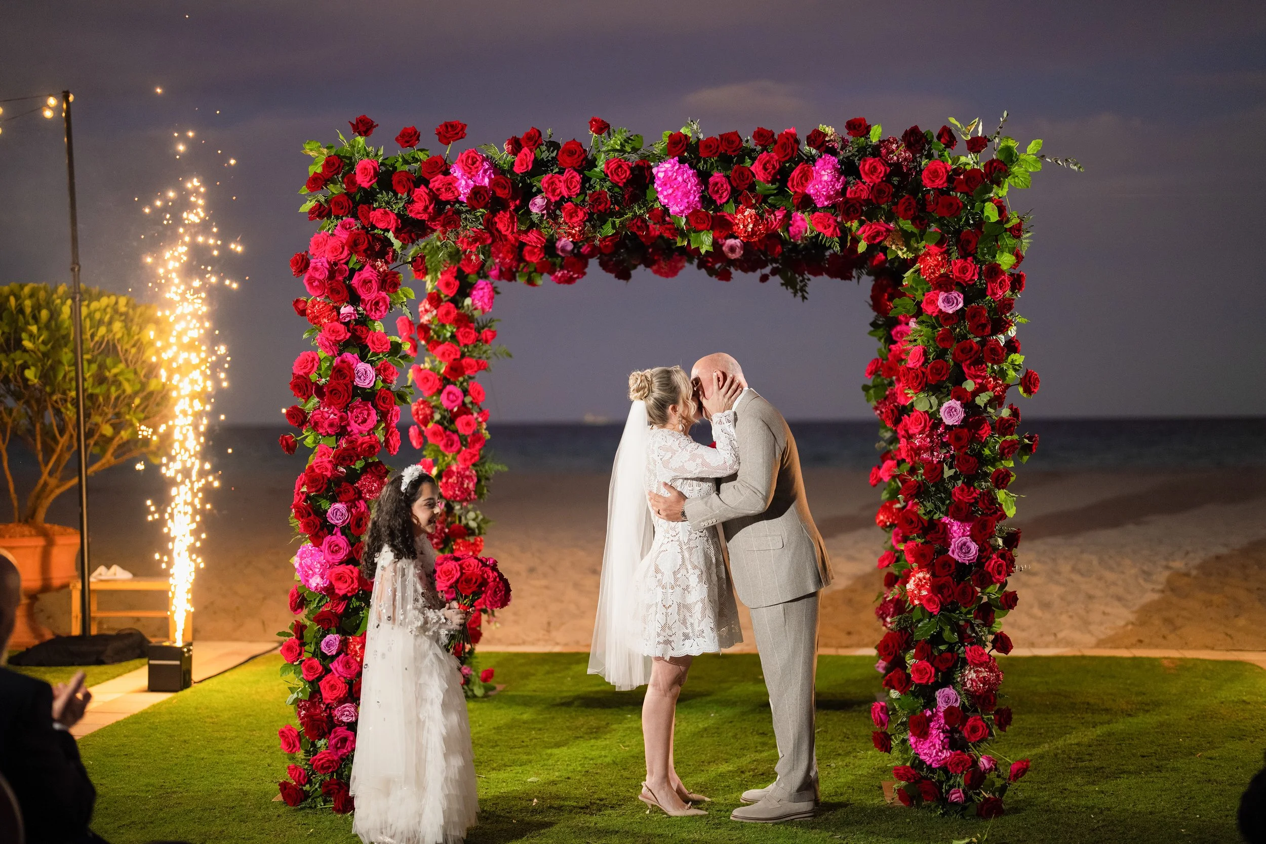 A wedding ceremony taking place outdoors at sunset, with a floral arch of red and pink roses, a bride and groom kissing, a young bridesmaid holding flowers, and fireworks in the background near the beach.