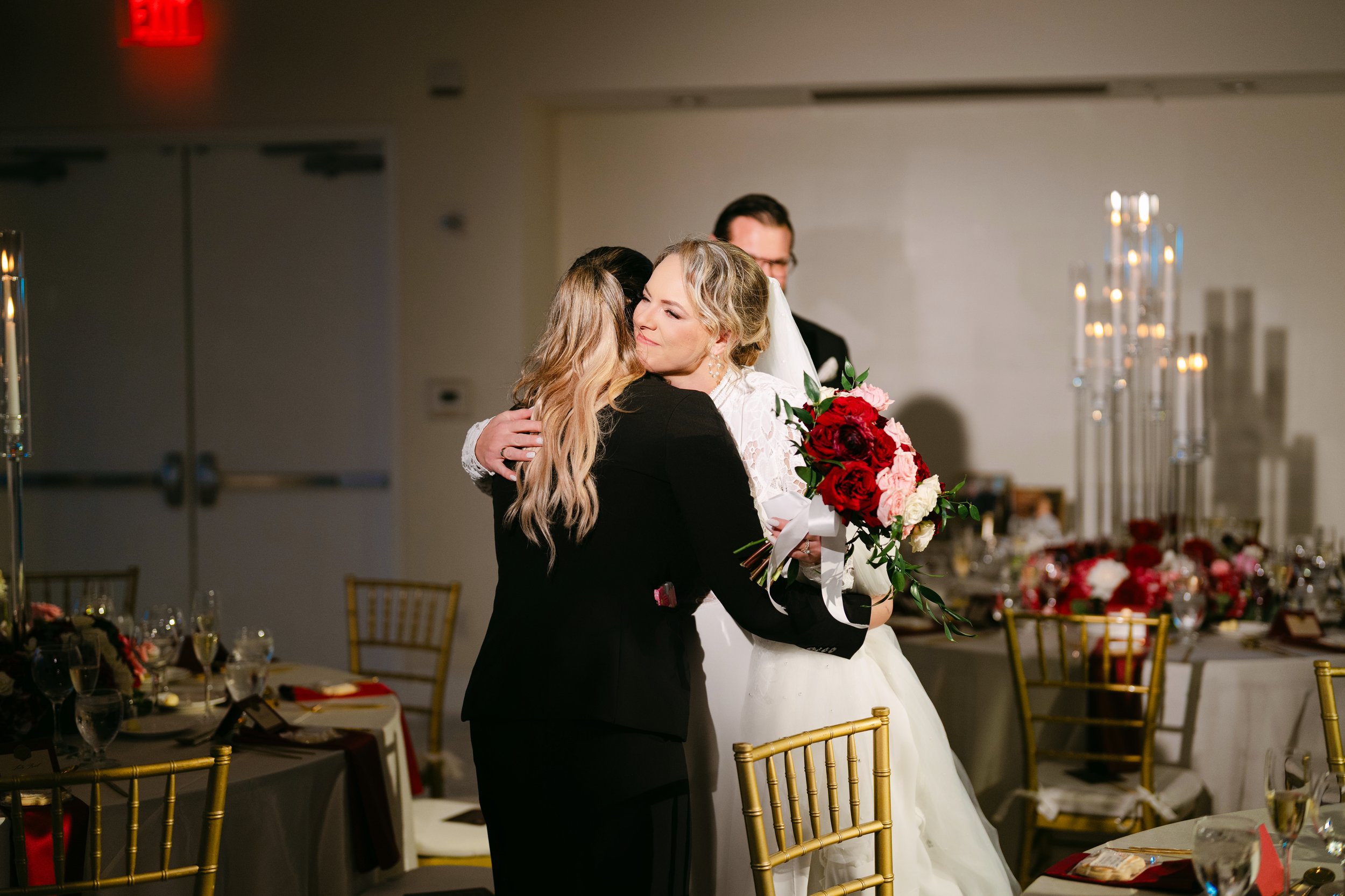 A bride receiving a warm hug at her wedding reception, holding a bouquet of red and white roses, in a decorated banquet hall with gold chairs and floral centerpieces.