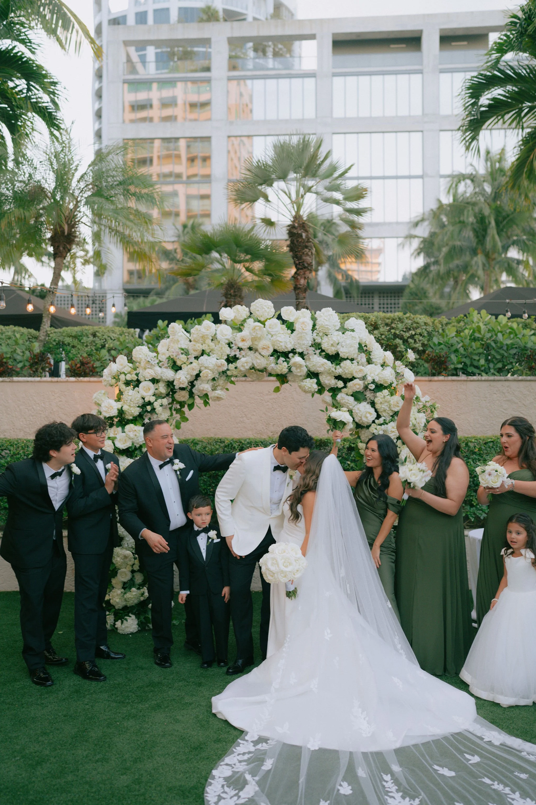 A newly married couple sharing a kiss under a floral wedding arch surrounded by friends and family, outdoors in a garden setting with palm trees and a modern building in the background.