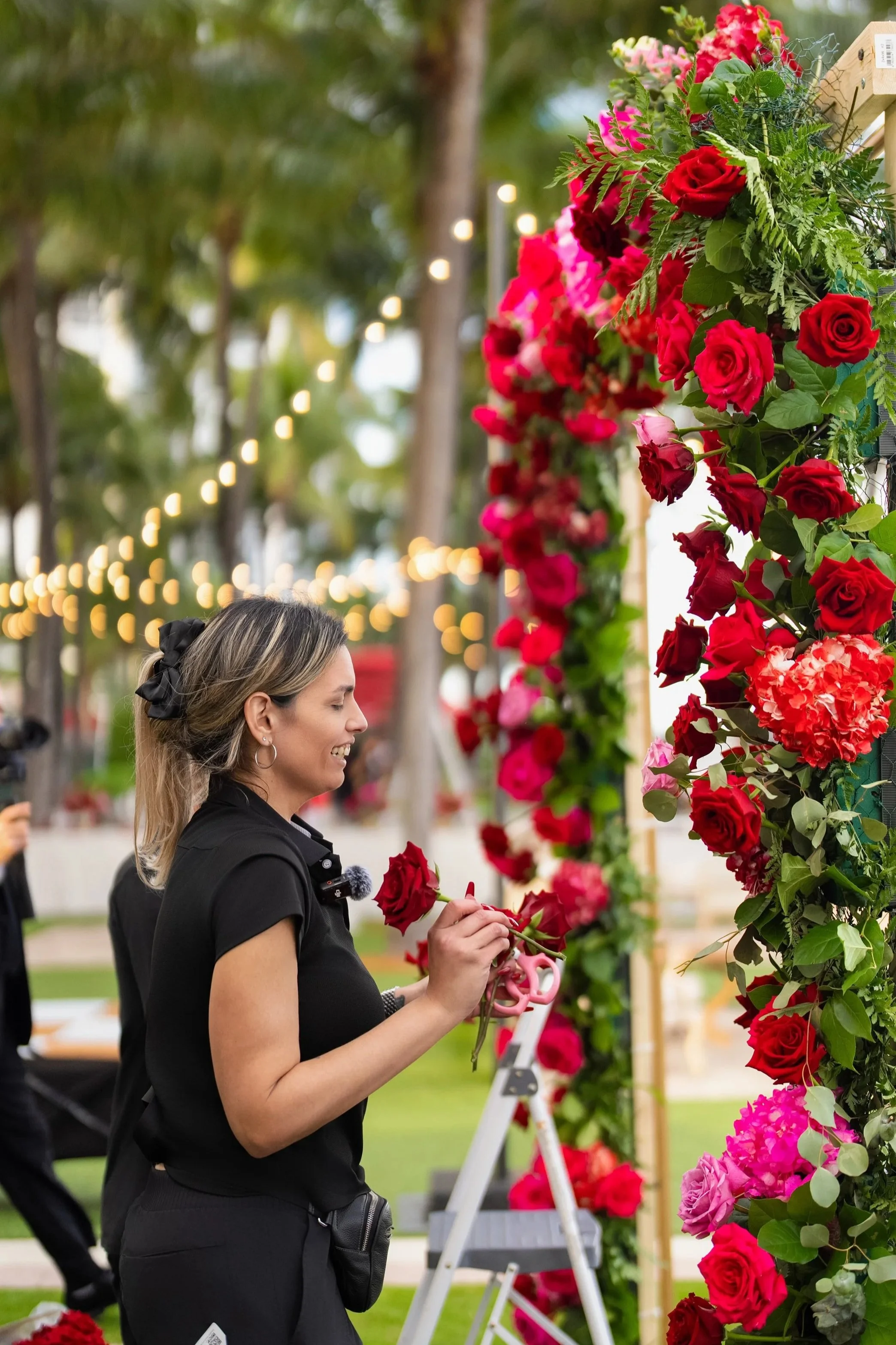 A woman arranging red and pink flowers on a decorative floral arch outdoors during the evening, with string lights overhead.