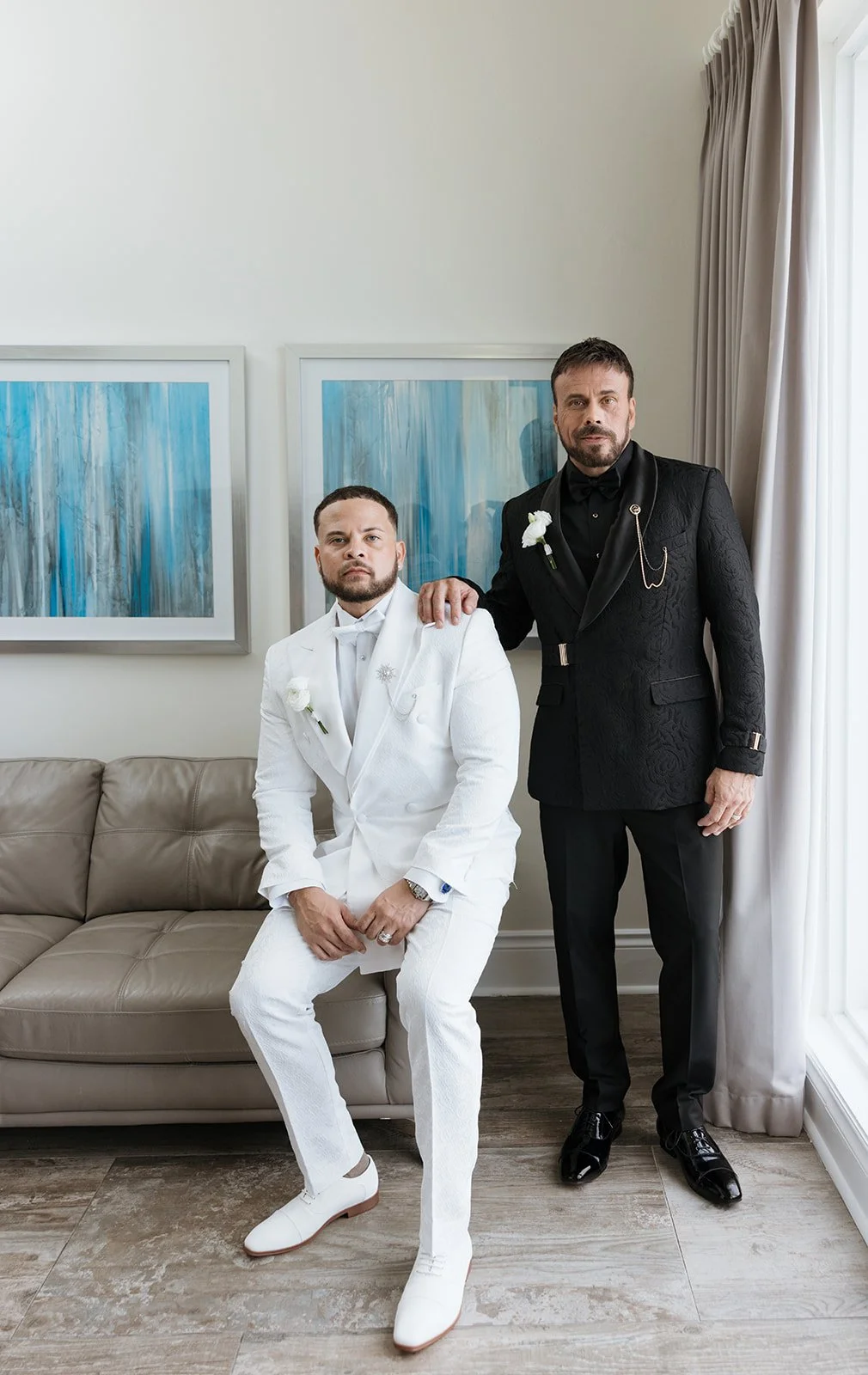 Two men in formal suits, one in a white tuxedo sitting on a beige couch and the other in a black tuxedo standing beside him with one hand on his shoulder, inside a room with blue artwork on the wall and white curtains.