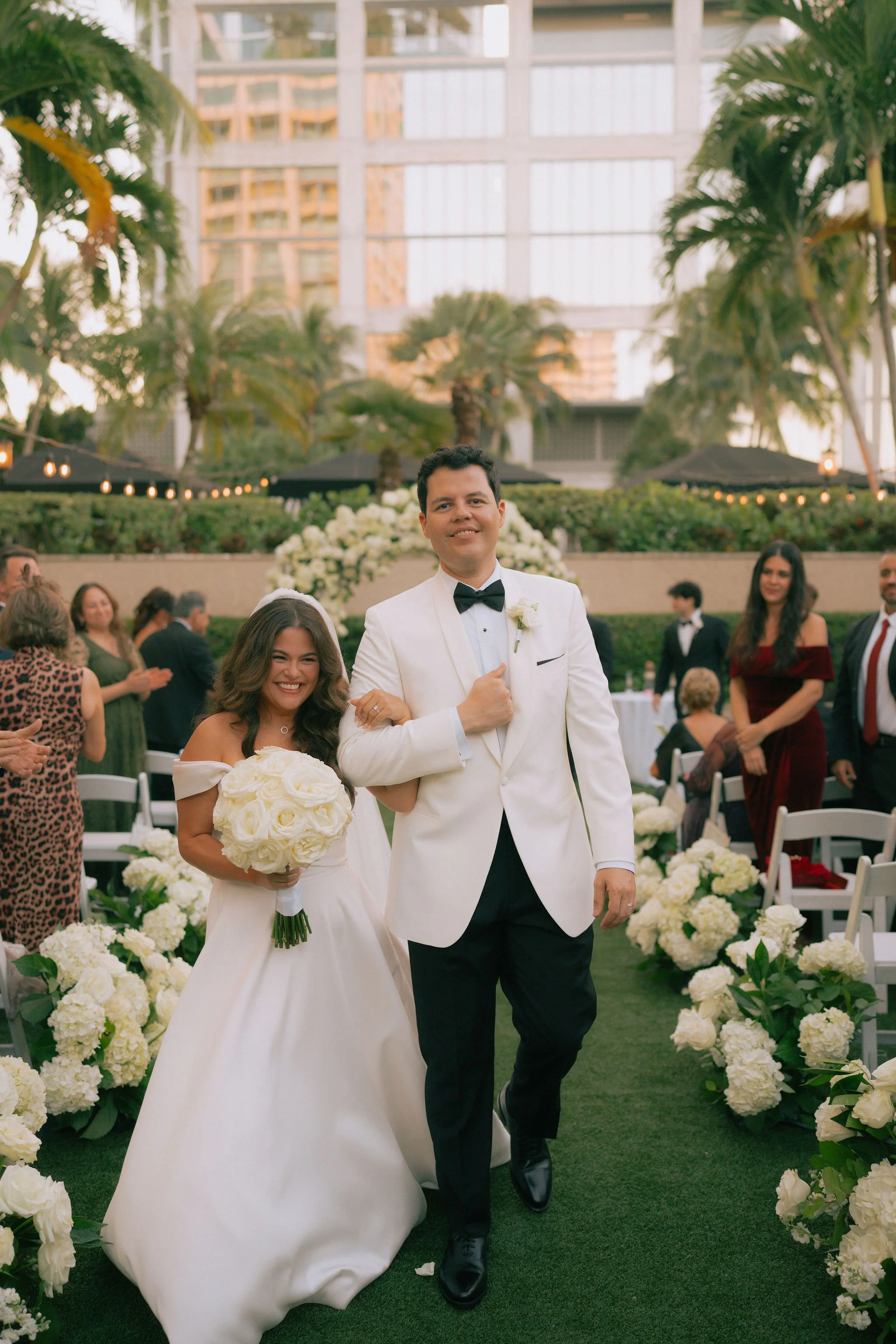 A couple dressed in wedding attire walking down an aisle decorated with white flowers with guests in the background at an outdoor wedding venue.