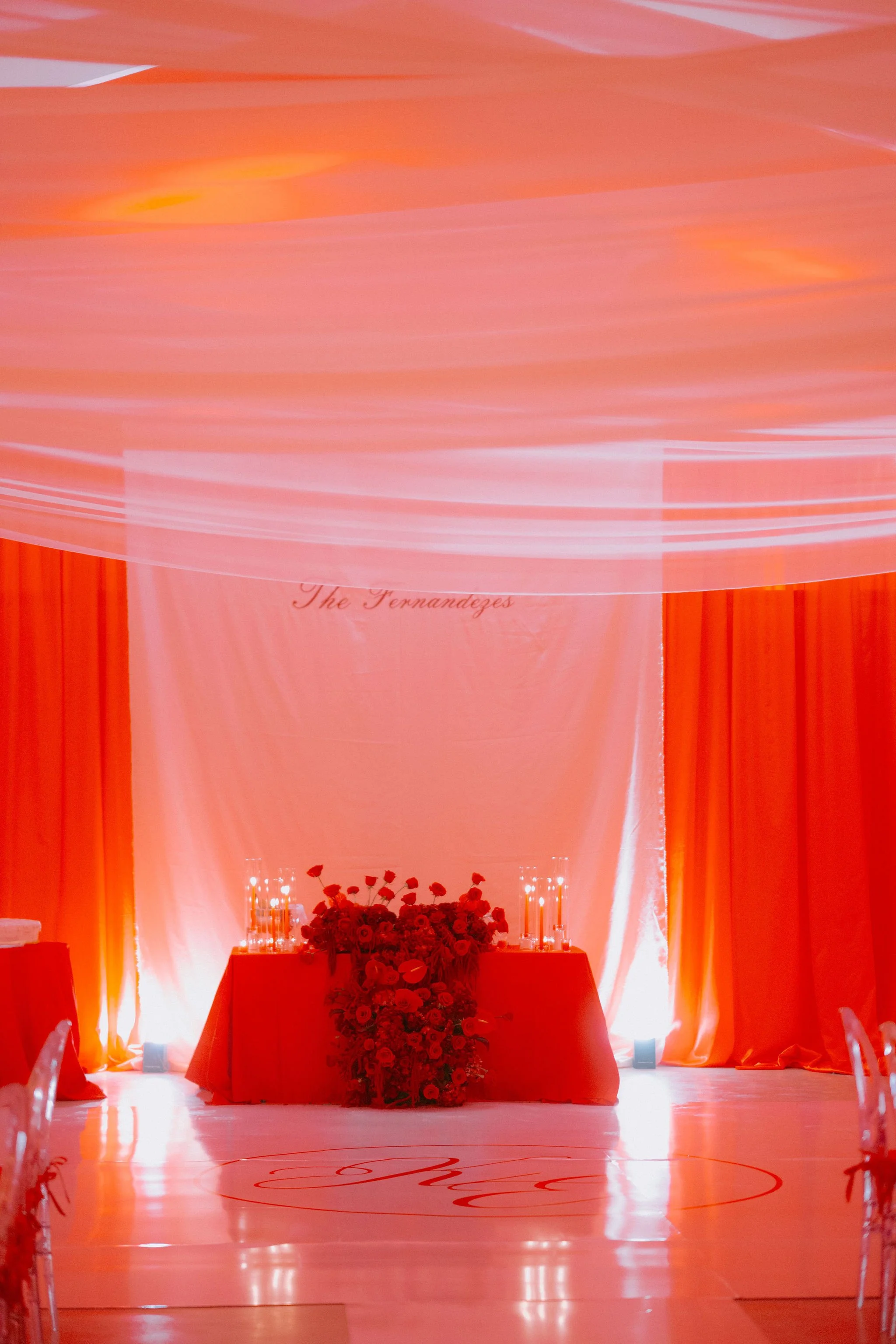 Decorated event space with orange lighting, draped curtains, and a table with red flowers and candles, likely set for a celebration or ceremony.