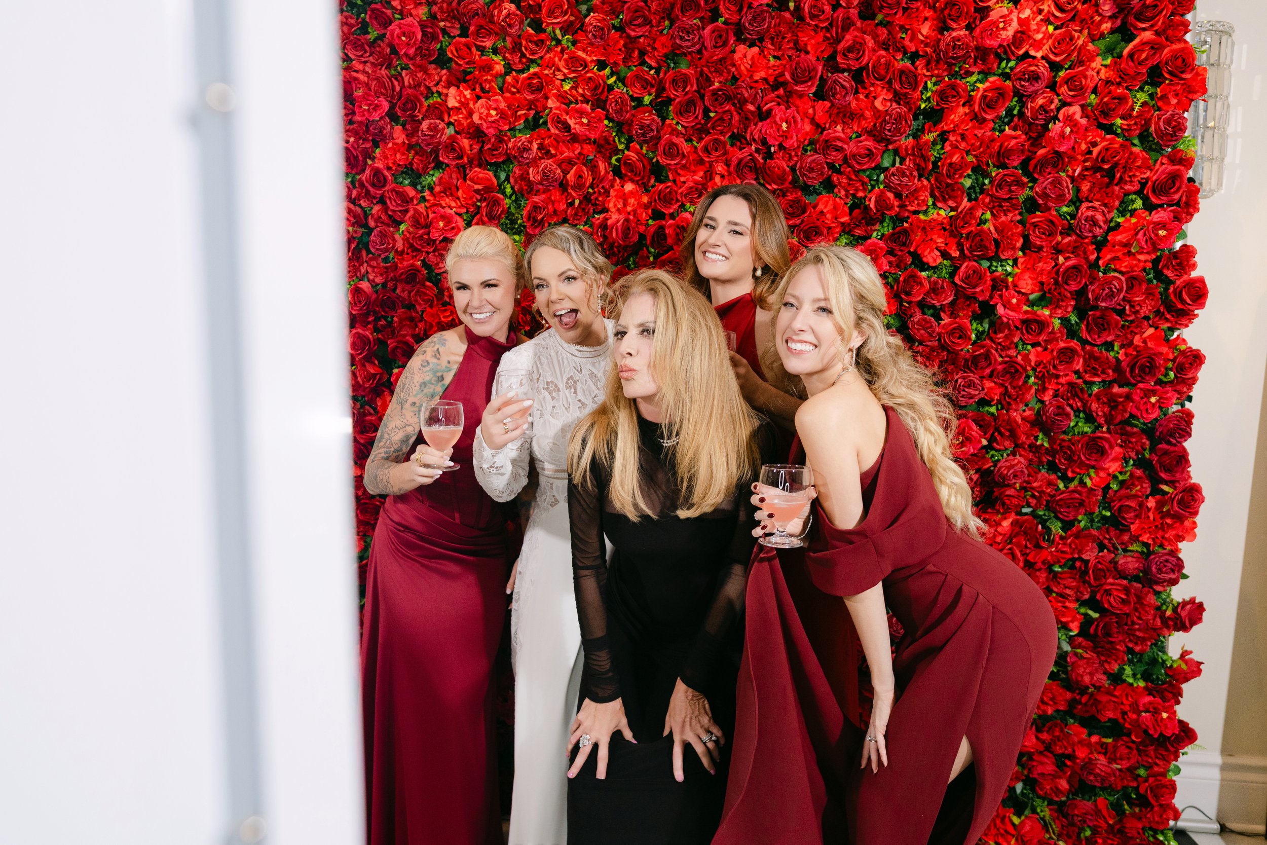 Group of six women in formal dresses taking a selfie in front of a red rose wall. Some are holding glasses of wine, and they are smiling or making playful faces.