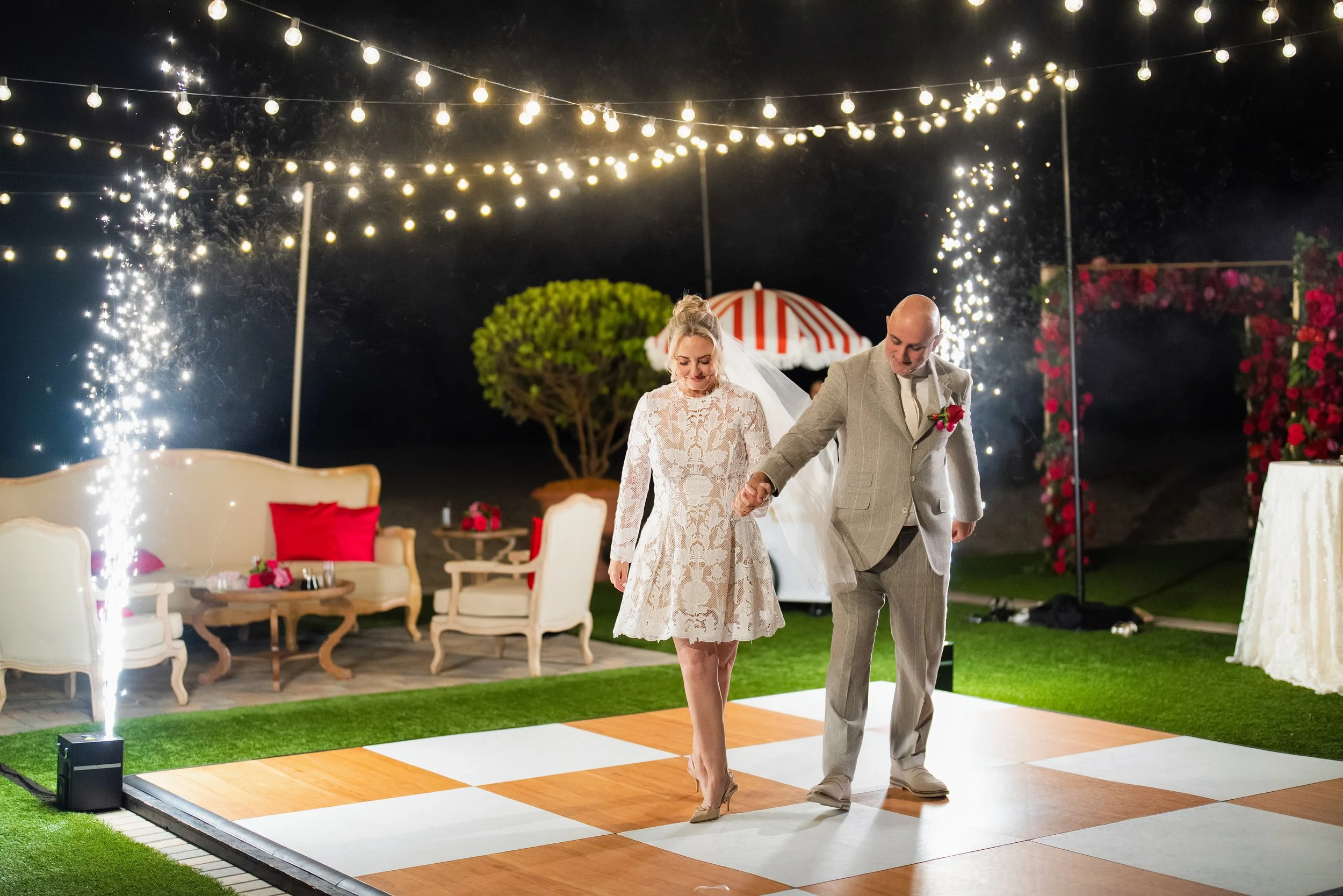 Bride and groom dancing on a checkered dance floor at night with string lights overhead, surrounded by outdoor furniture and a fireworks display