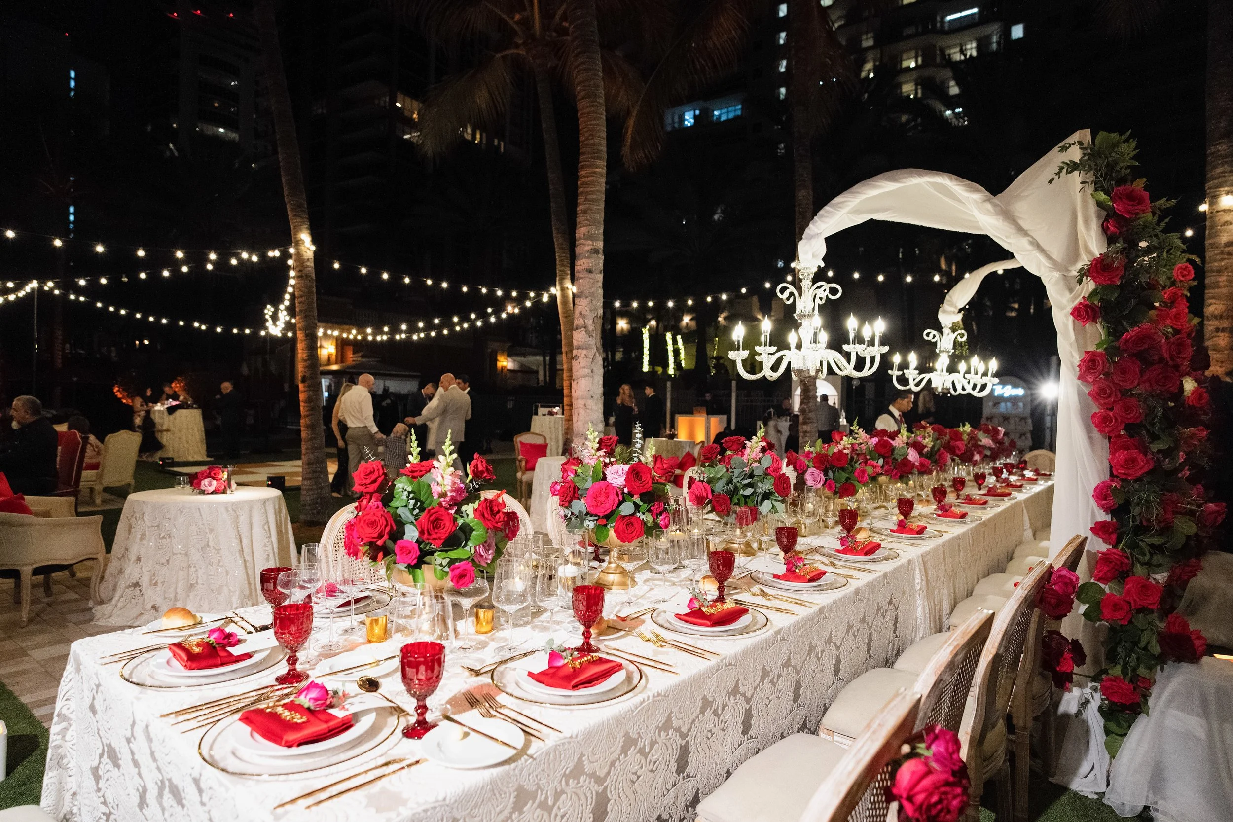 A long banquet table decorated with pink and red roses, gold cutlery, red glassware, and red napkins, set for an elegant outdoor event at night with string lights, palm trees, and chandeliers in the background.