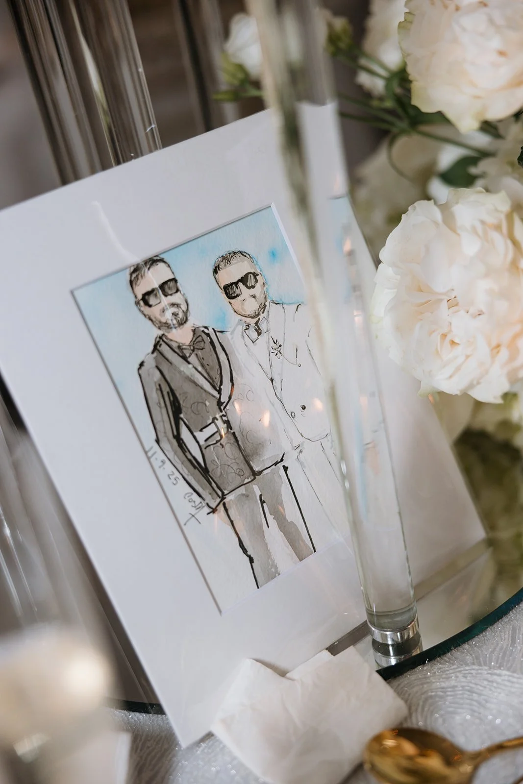A hand-drawn illustration of two men wearing suits and sunglasses, framed and placed on a table with white flowers and a glass object nearby.