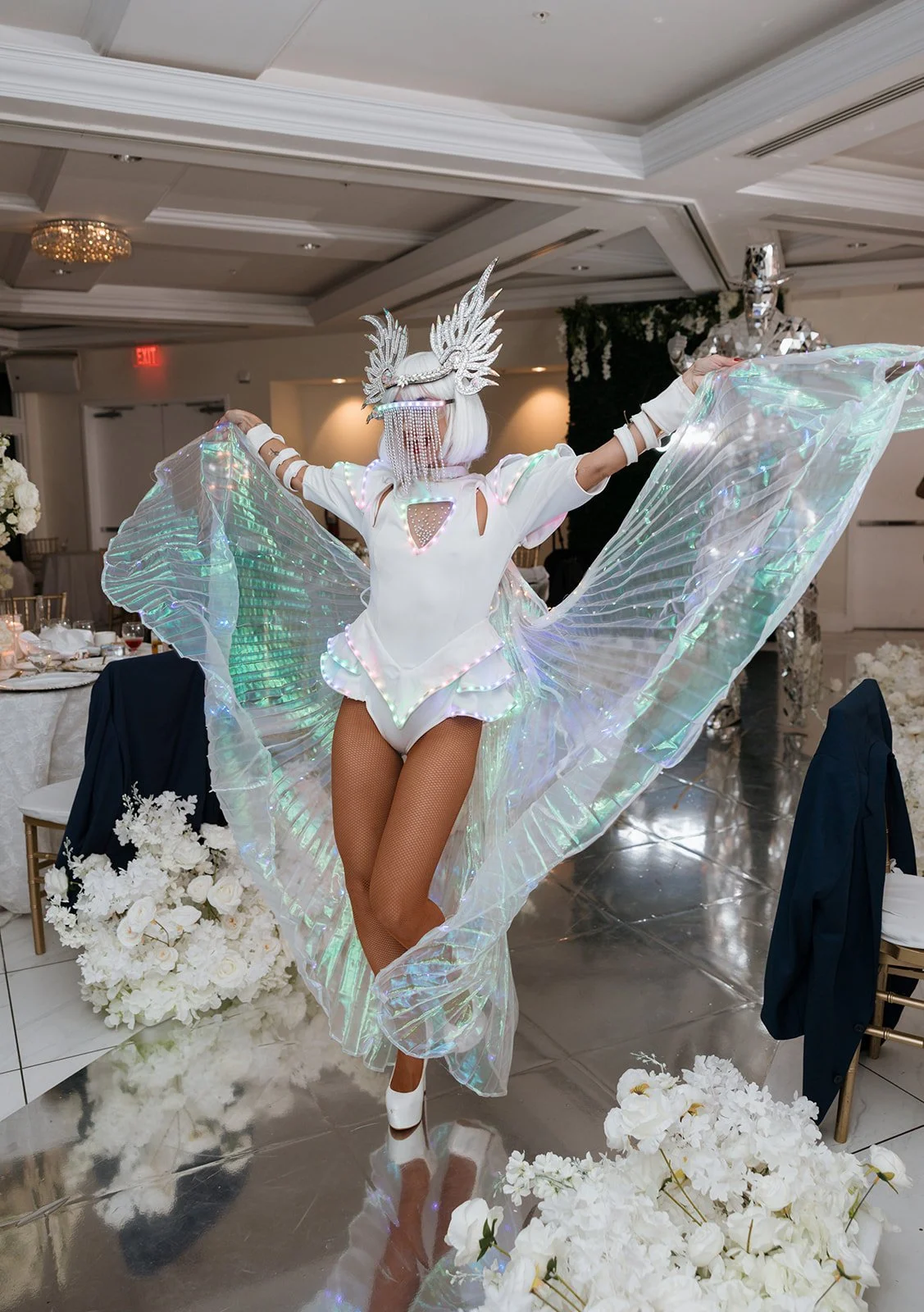 A performer in an elaborate, futuristic costume with iridescent wings and a silver headdress, posing at an event with decorated tables and floral arrangements.