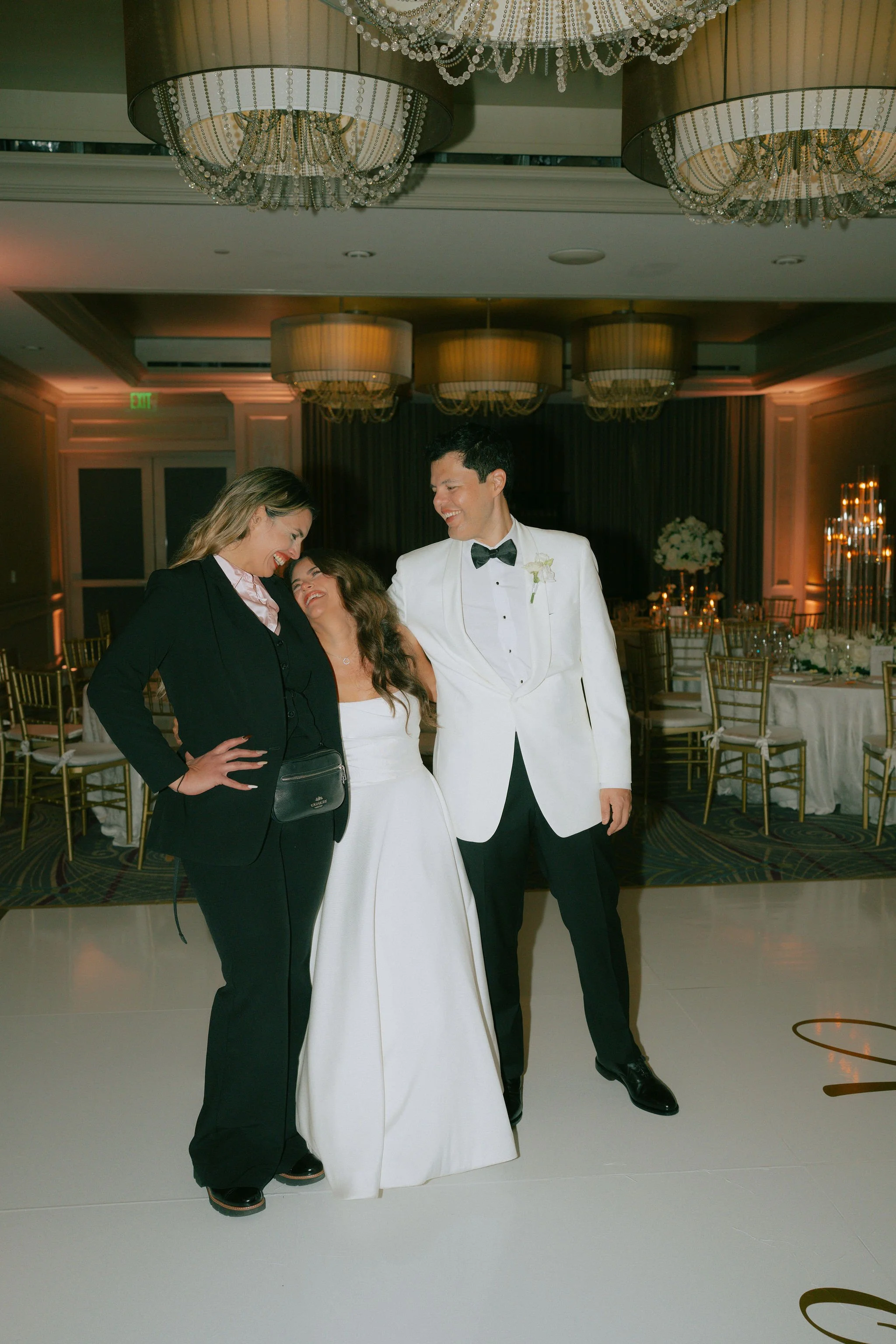 A bride and groom standing with a woman at a wedding reception, smiling and enjoying each other's company, in an elegant banquet hall with chandeliers and decorated tables.