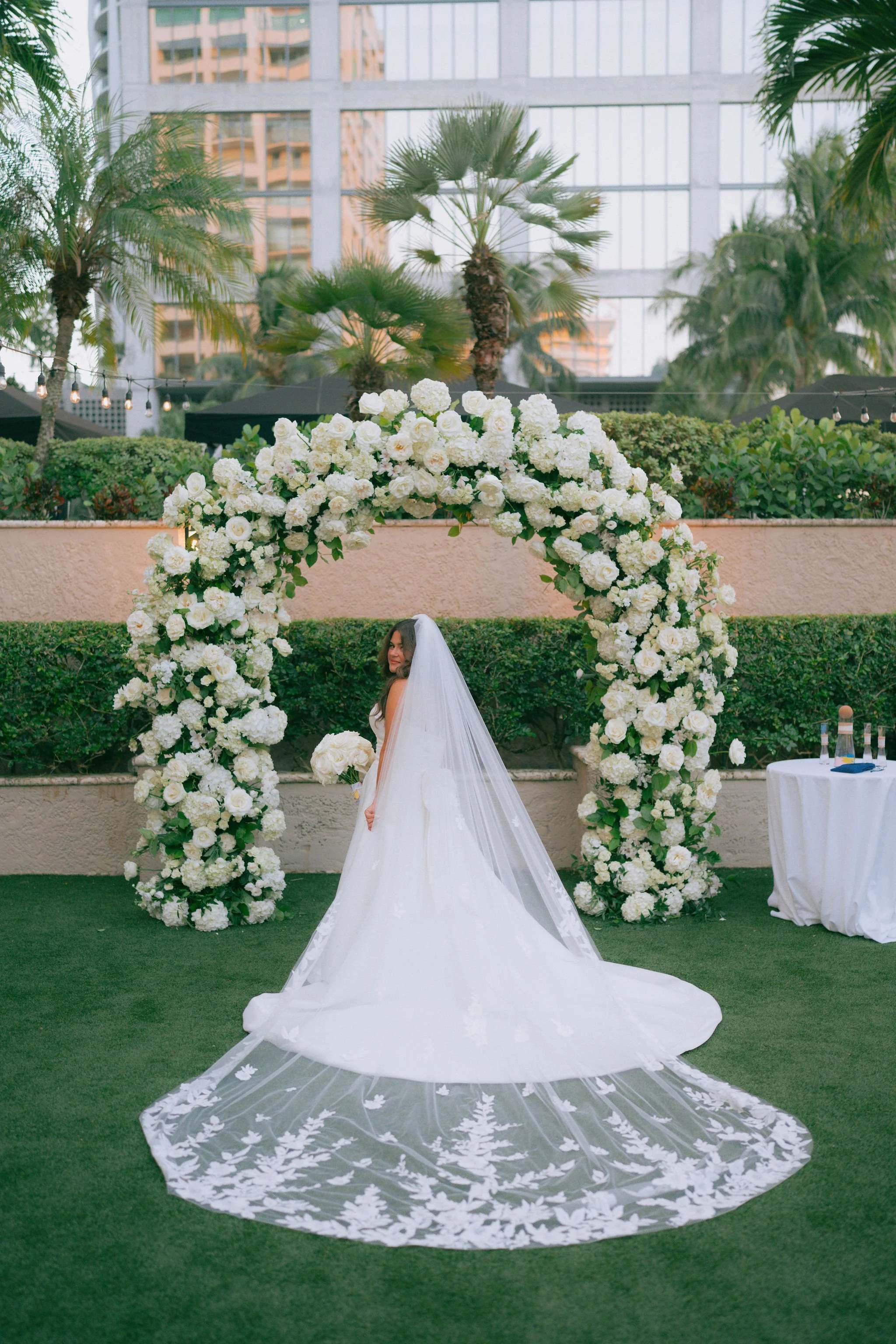 A bride in a white wedding gown and veil stands outdoors under a floral arch with white roses, surrounded by greenery and palm trees, with an urban backdrop of tall buildings.