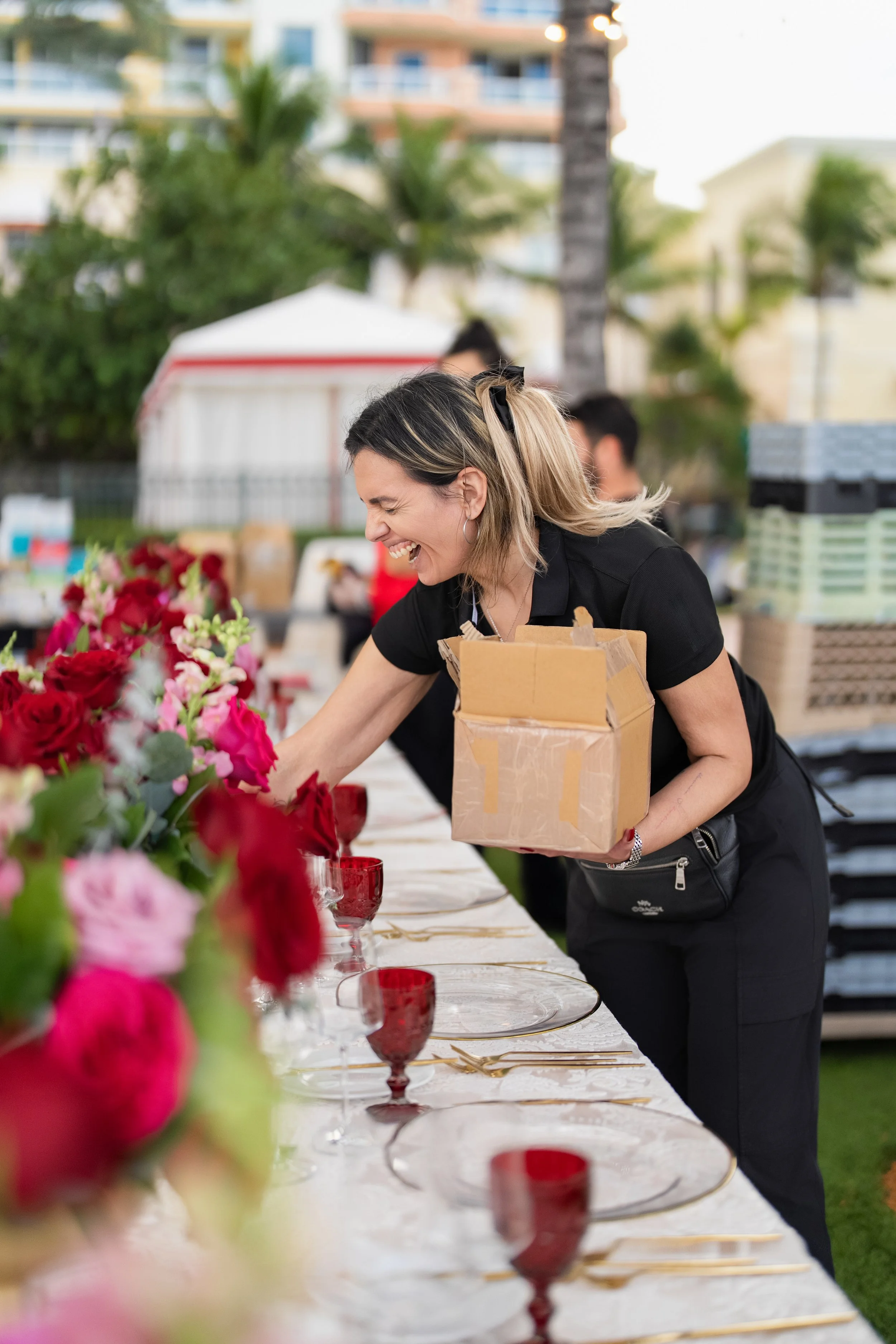 Woman arranging flowers on a table outdoors, smiling, with a cardboard box in her hand, flowers and glassware on the table, palm trees, and buildings in the background.