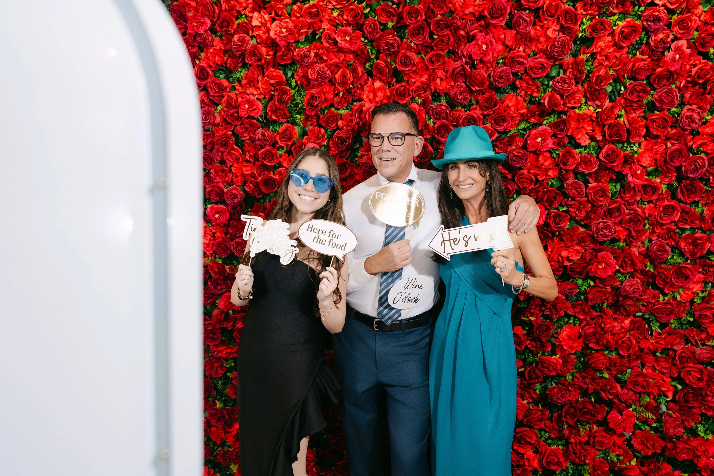 Three adults at a photo booth with a backdrop of red roses, holding signs and wearing playful accessories, smiling at the camera.