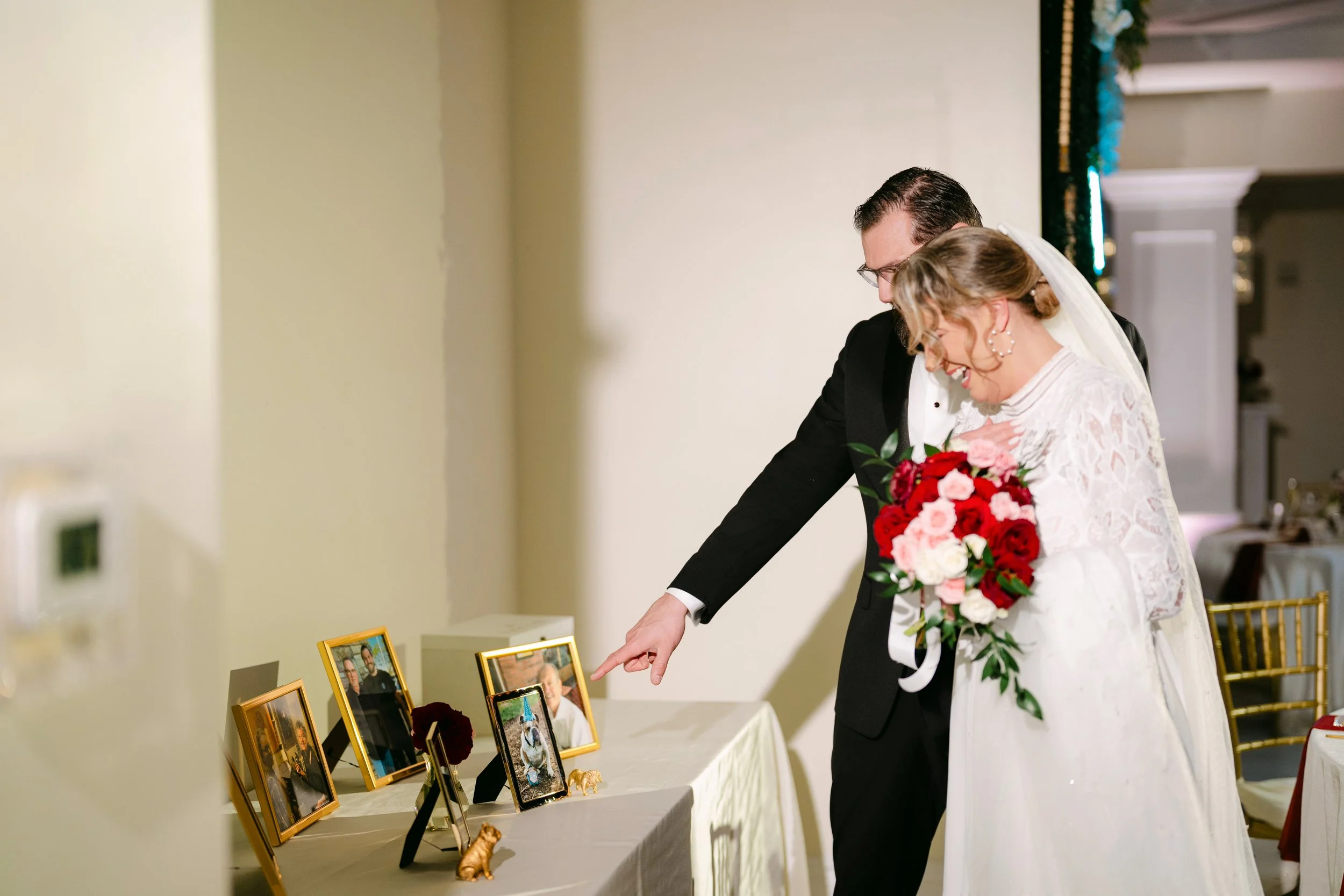 A bride and groom at a wedding reception looking at framed photographs on a table. The bride holds a bouquet of red and pink flowers, and the groom points at one of the photograph frames.