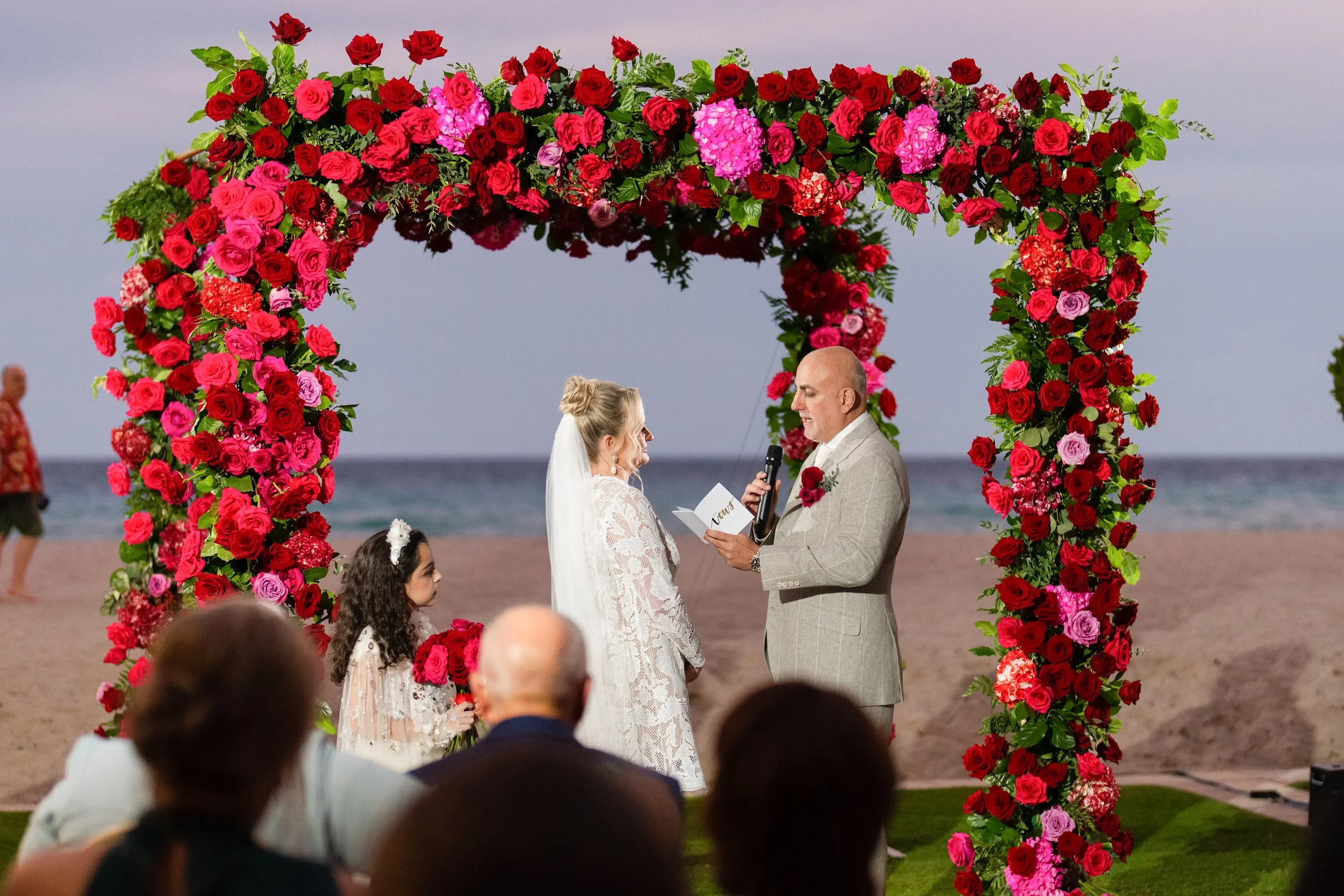 A couple getting married on a beach, exchanging vows under a large floral arch made of pink and red roses with greenery, with the ocean in the background.