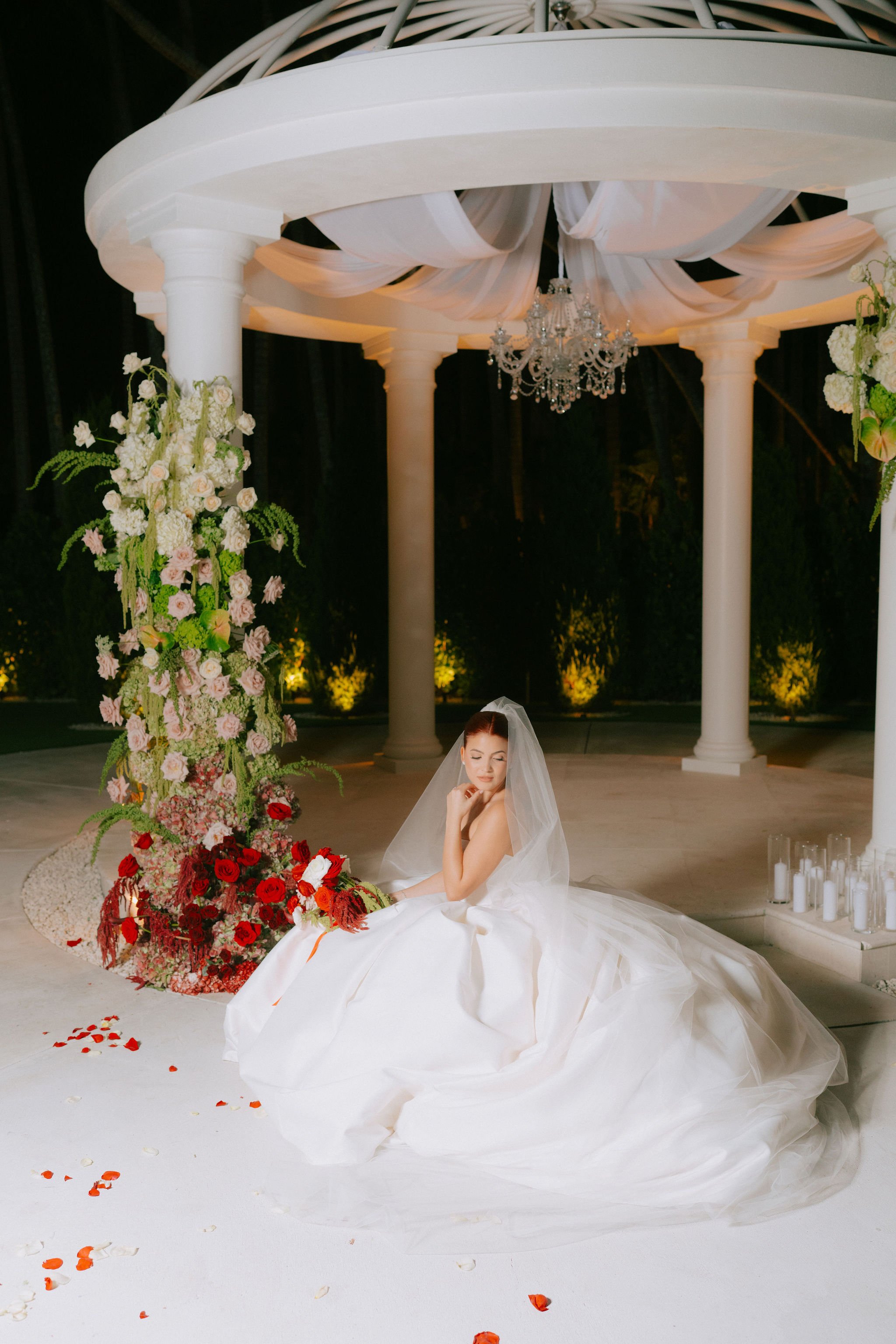 A bride in a white wedding gown and veil sitting on the floor next to a tall floral arrangement with pink, white, and red flowers under a white canopy with columns.
