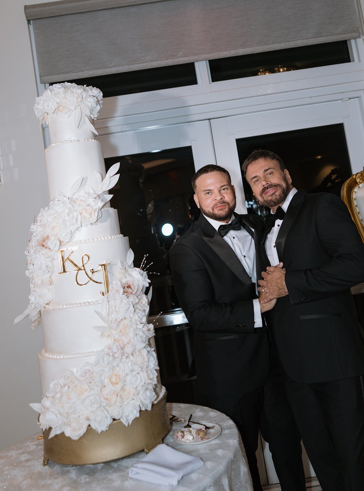 Two men in tuxedos holding hands, standing next to a large wedding cake decorated with white flowers and gold accents, with the initials "K & J" on the cake, inside a banquet hall with large windows in the background.