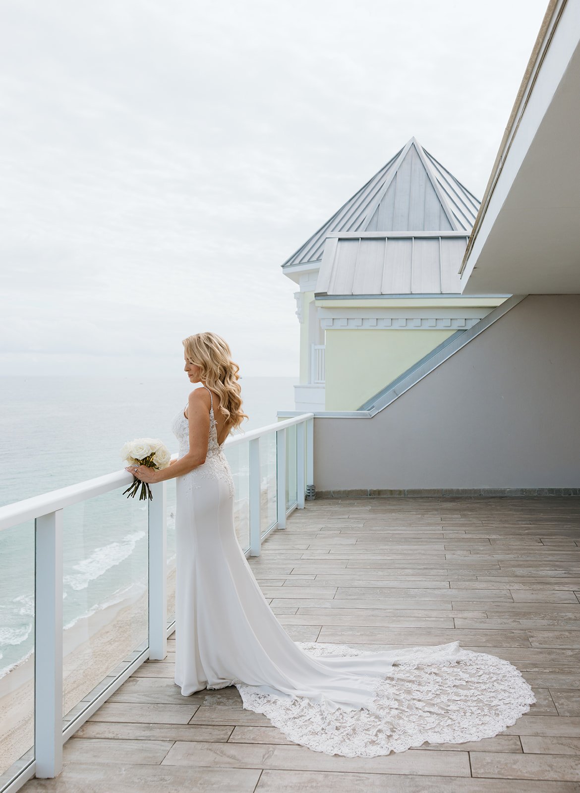 Bride in a white wedding gown holding a bouquet, standing on a balcony overlooking the ocean, with a white building and cloudy sky in the background.