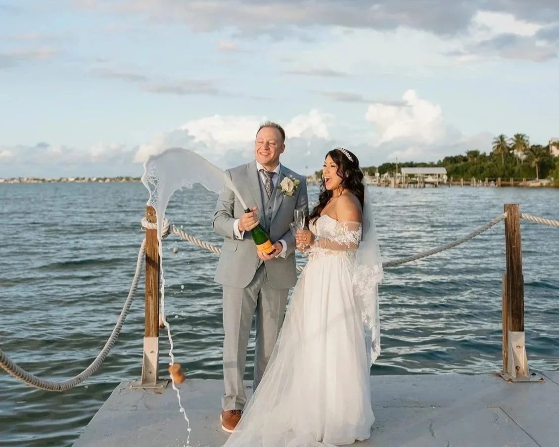 A newlywed couple celebrating on a dock by the water. The groom in a gray suit and the bride in a white wedding dress are popping a bottle of champagne, with the champagne foam spouting out. They are smiling and holding glasses, with a scenic water and sky background.