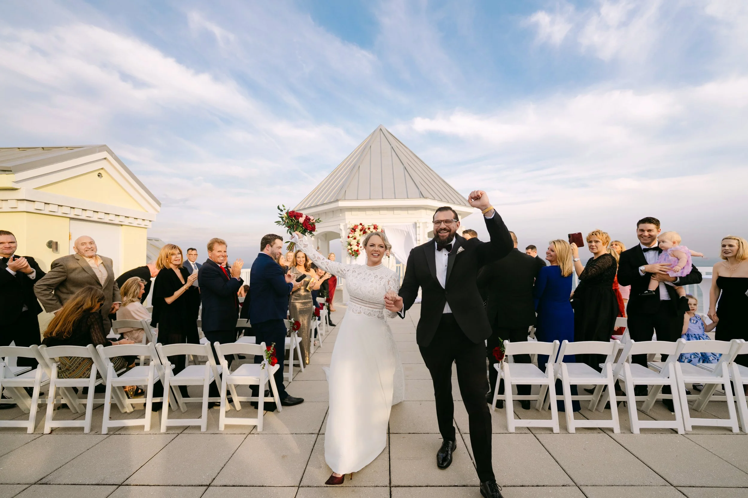 A wedding ceremony with an officiant speaking into a microphone, a bride and groom holding hands in front of an arch decorated with greenery and white drapes.