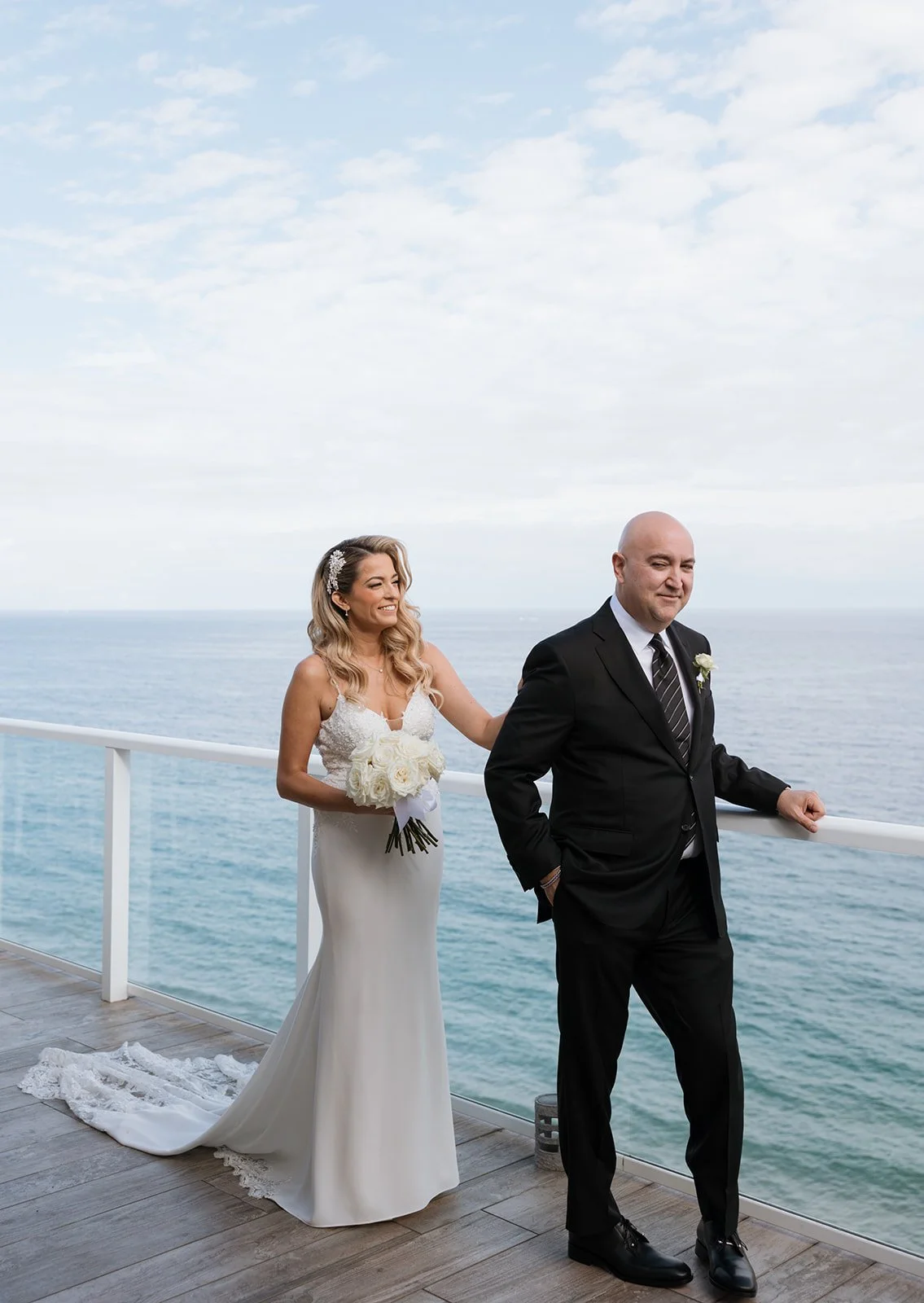 A bride and groom standing on a balcony overlooking the ocean during their wedding. The bride is wearing a white wedding dress and holding a bouquet of white roses, while the groom is dressed in a black suit and striped tie, leaning against the raili