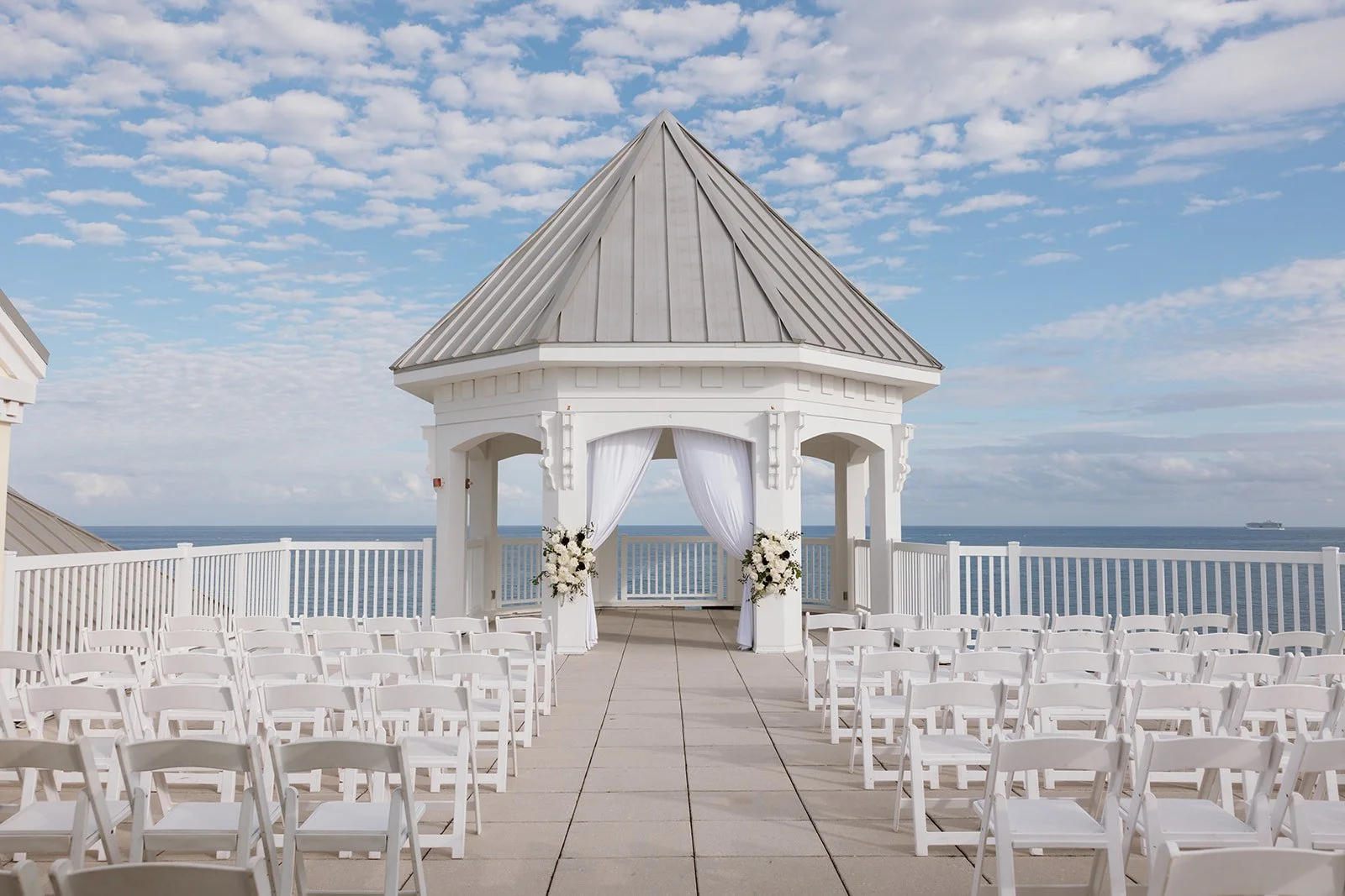 White outdoor wedding gazebo decorated with flowers overlooking the ocean under a partly cloudy sky.