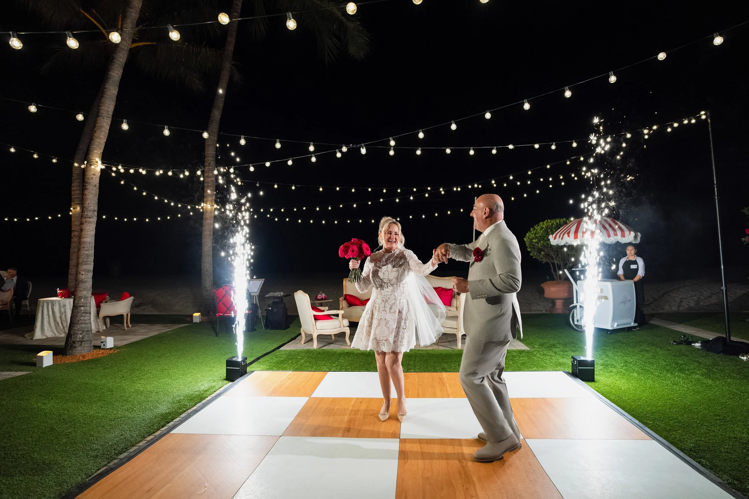 A bride and groom dancing on a wedding dance floor at night under string lights, with fireworks and a DJ in the background.