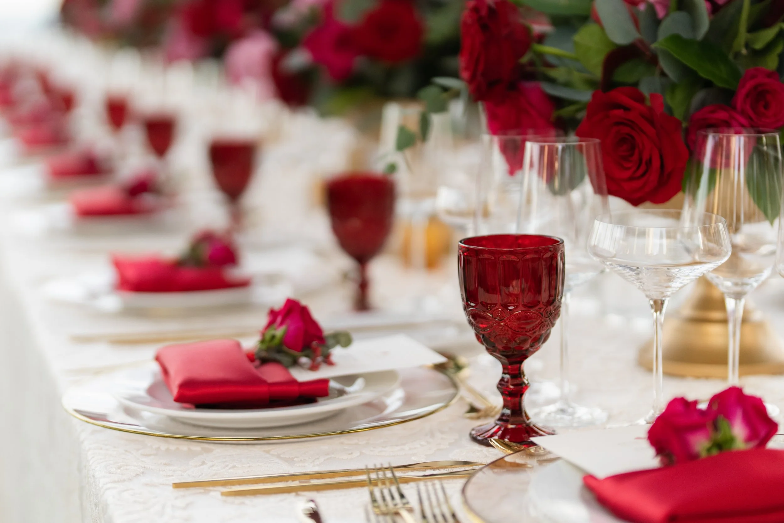Elegant dining table set with red and pink roses, red and clear glassware, gold-accented plates, and pink napkins.