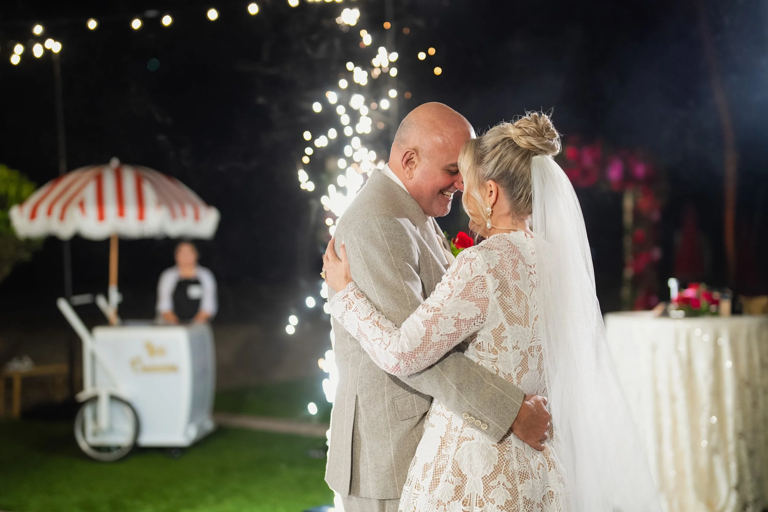 A bride and groom share a dance at their wedding reception outdoors at night, with sparklers and string lights in the background.