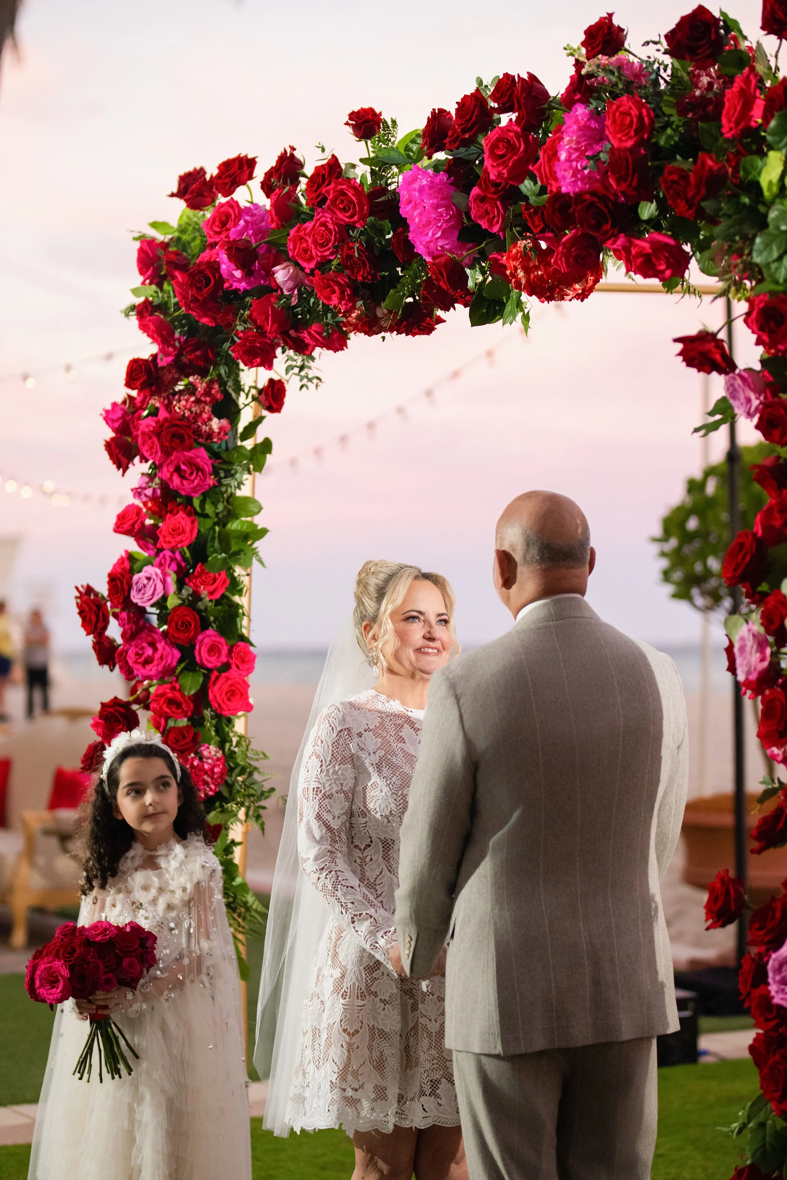 A bride and groom holding hands under a big floral arch during their wedding ceremony on the beach at sunset, with a young girl holding a bouquet of pink roses standing nearby.