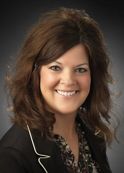 Professional woman with shoulder-length brown hair, smiling, wearing a black blazer with a patterned blouse, against a dark background.